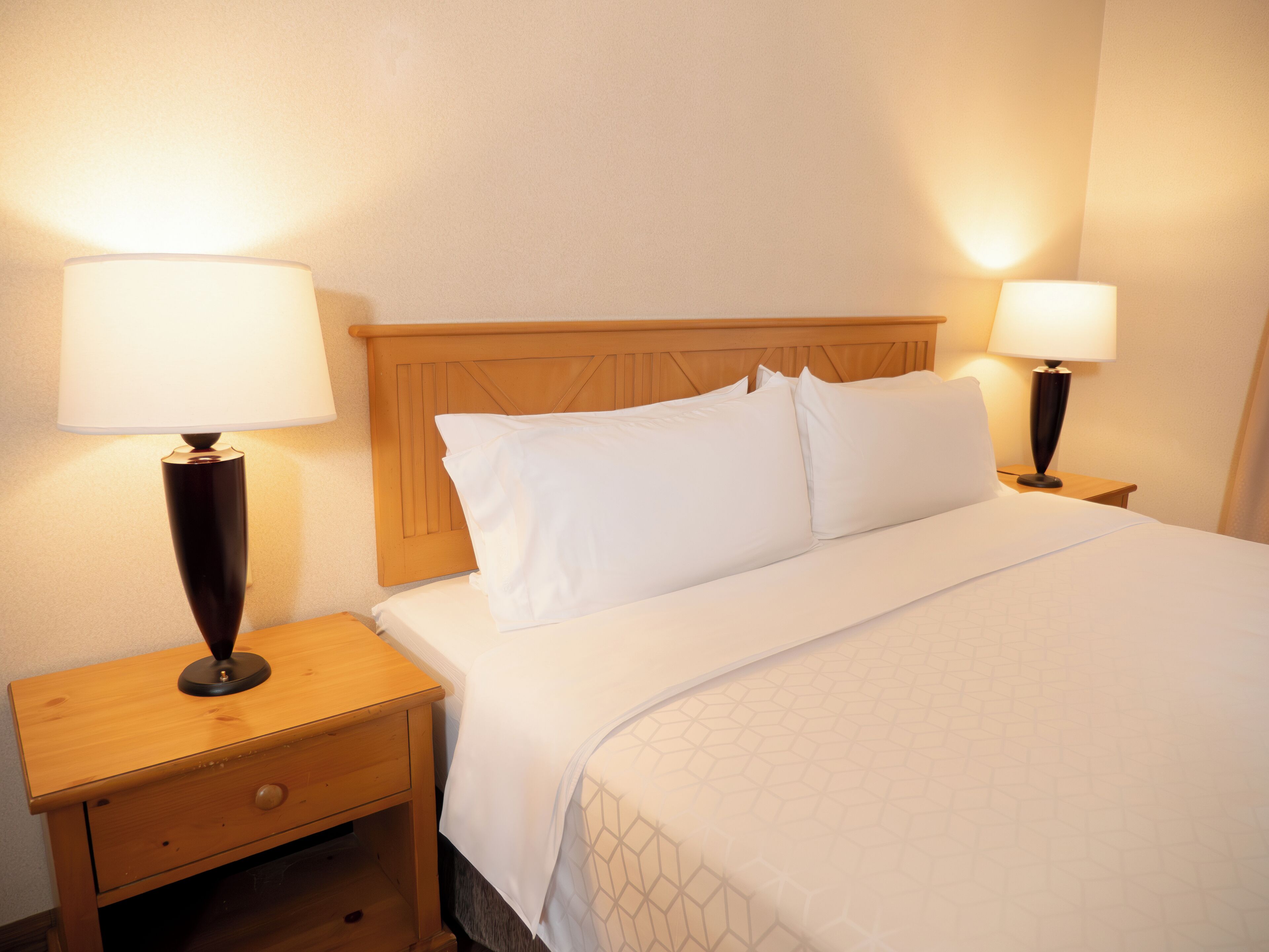 A neatly made bed with white linens, two bedside lamps, a wooden nightstand with a drawer, and a wooden headboard.