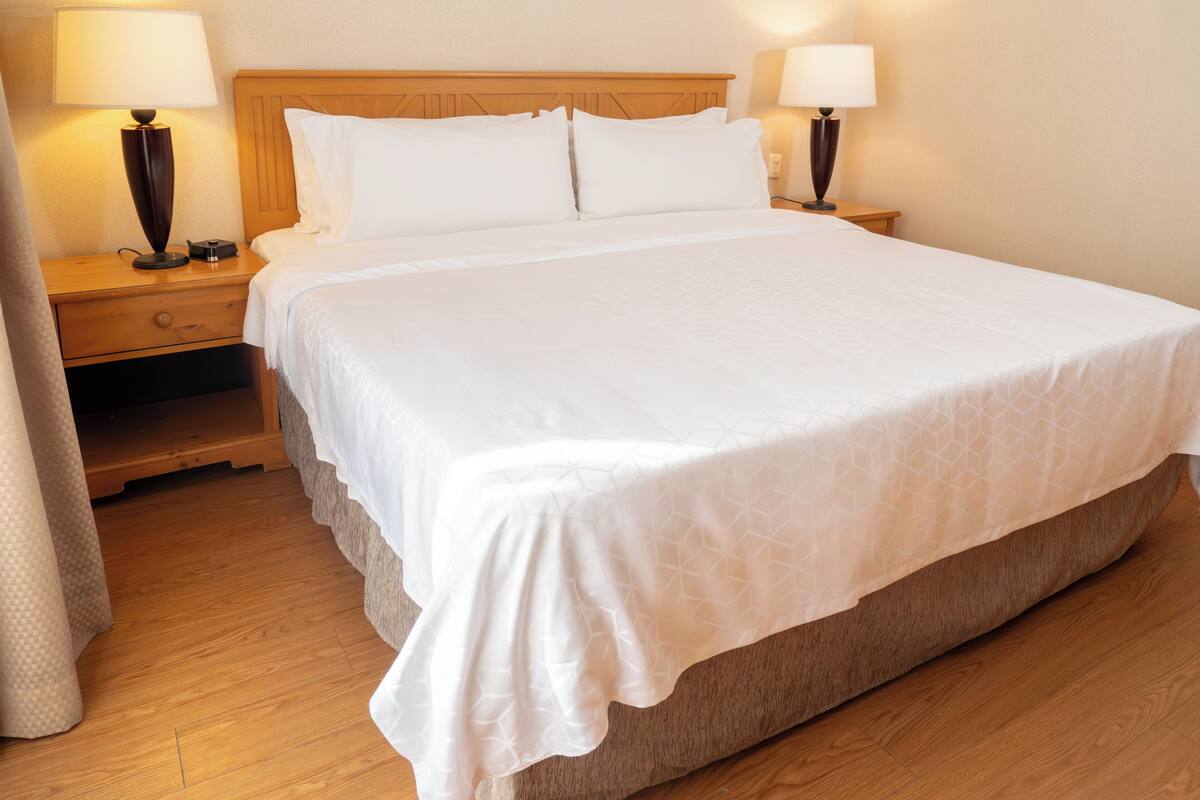 Una cama bien hecha con cabecera de madera, dos mesitas de noche con lámparas y suelo de madera.