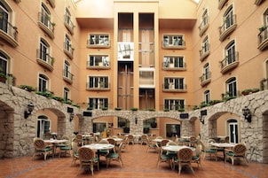 Un vestíbulo de hotel con varias mesas y sillas de comedor, una escalera central y balcones con plantas.