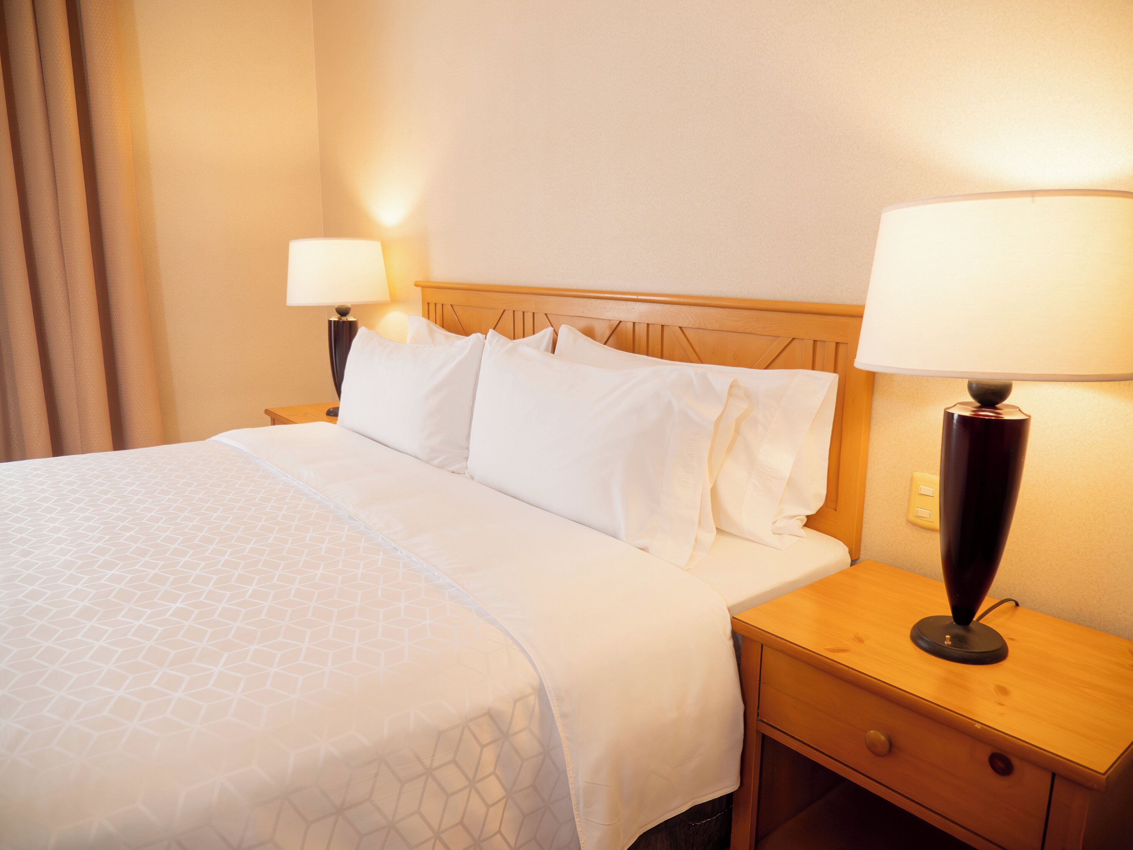 A hotel room with a neatly made bed, a wooden headboard, a nightstand with a lamp, and a wall socket.