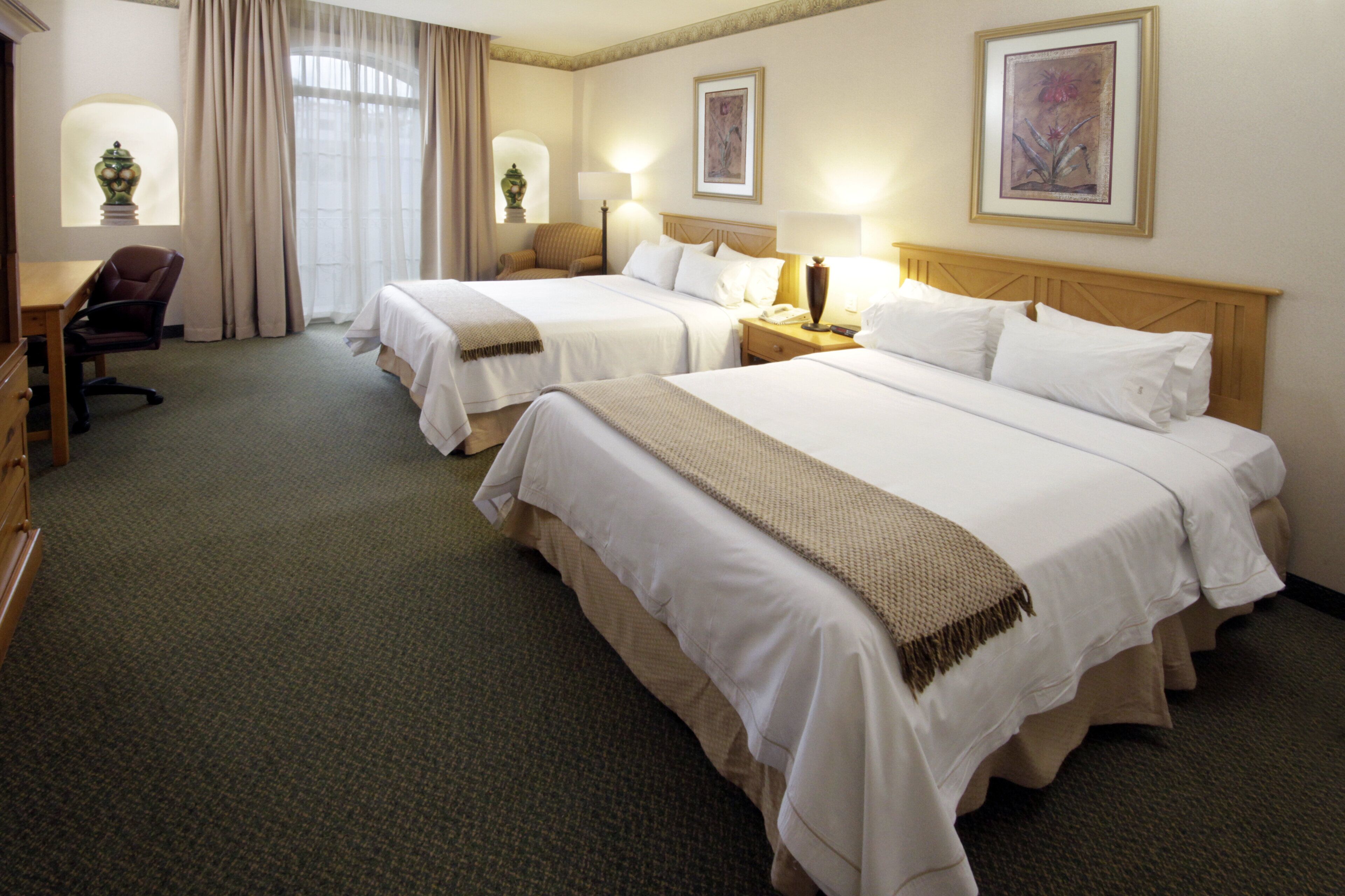 Habitación de hotel con dos camas, un escritorio, una silla y una ventana con cortinas.