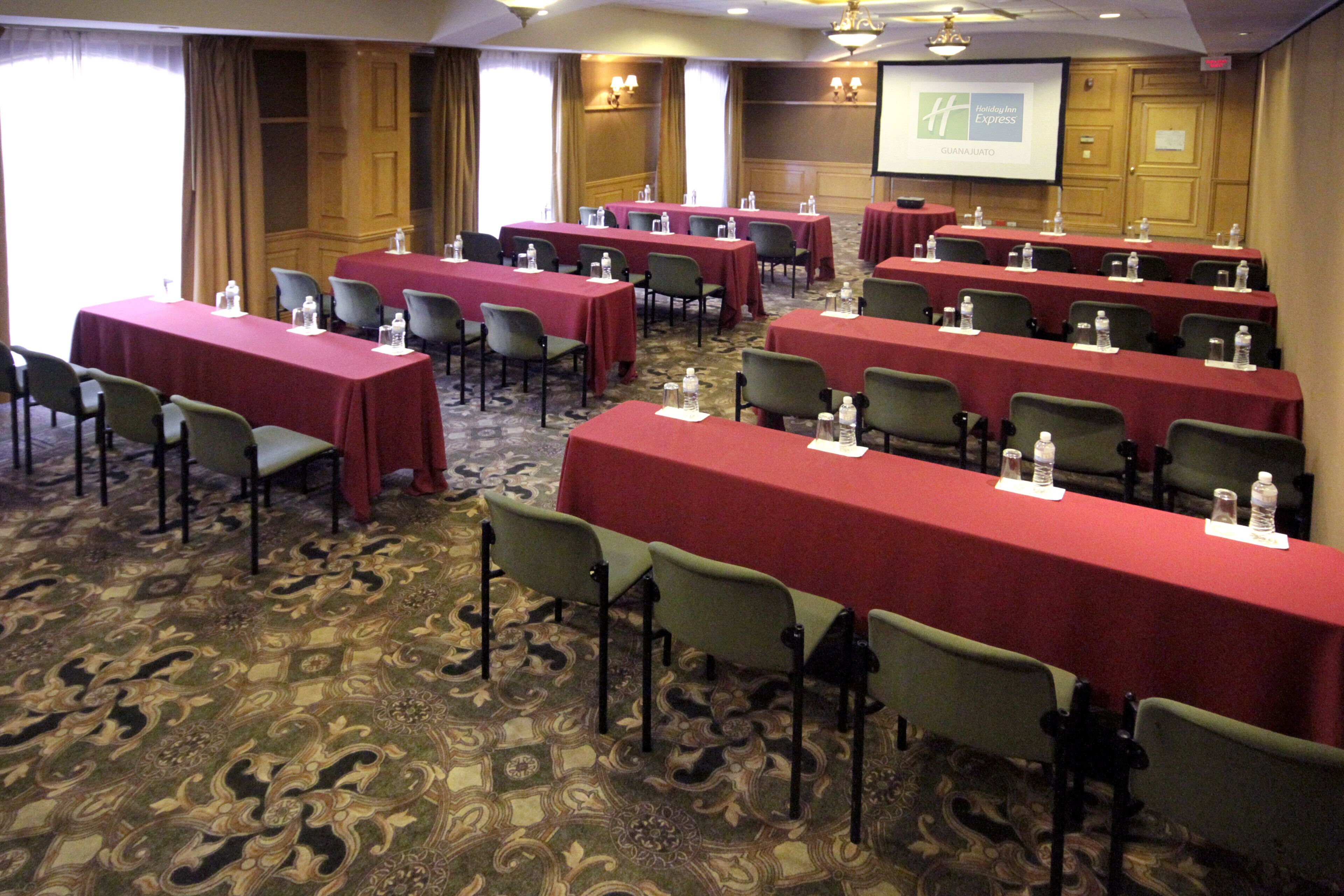 A conference room with multiple tables and chairs, arranged in rows, with a carpeted floor and a screen displaying the Holiday Inn Express logo.
