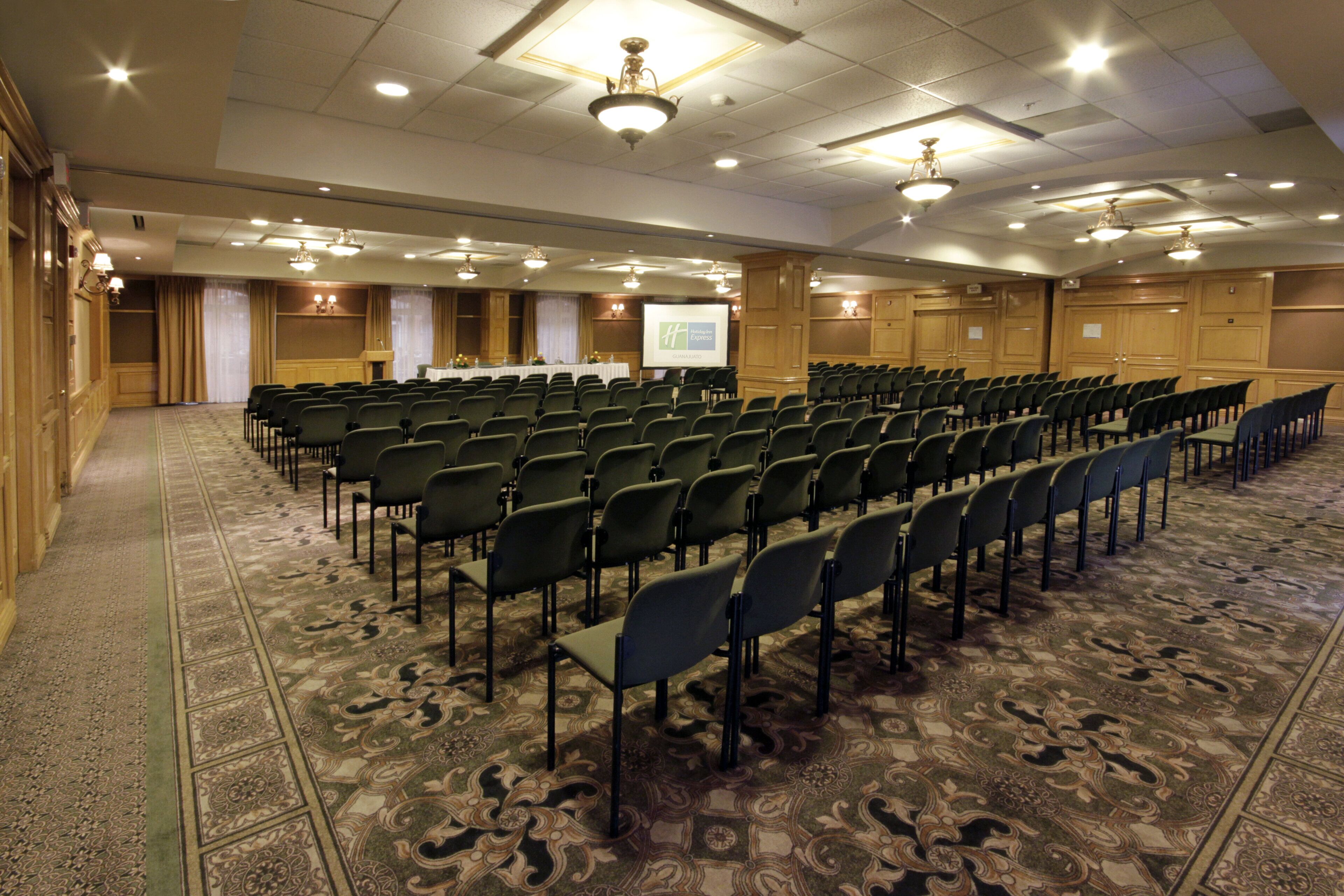 Un salón de conferencias con filas de sillas, un atril y una pantalla de presentación.