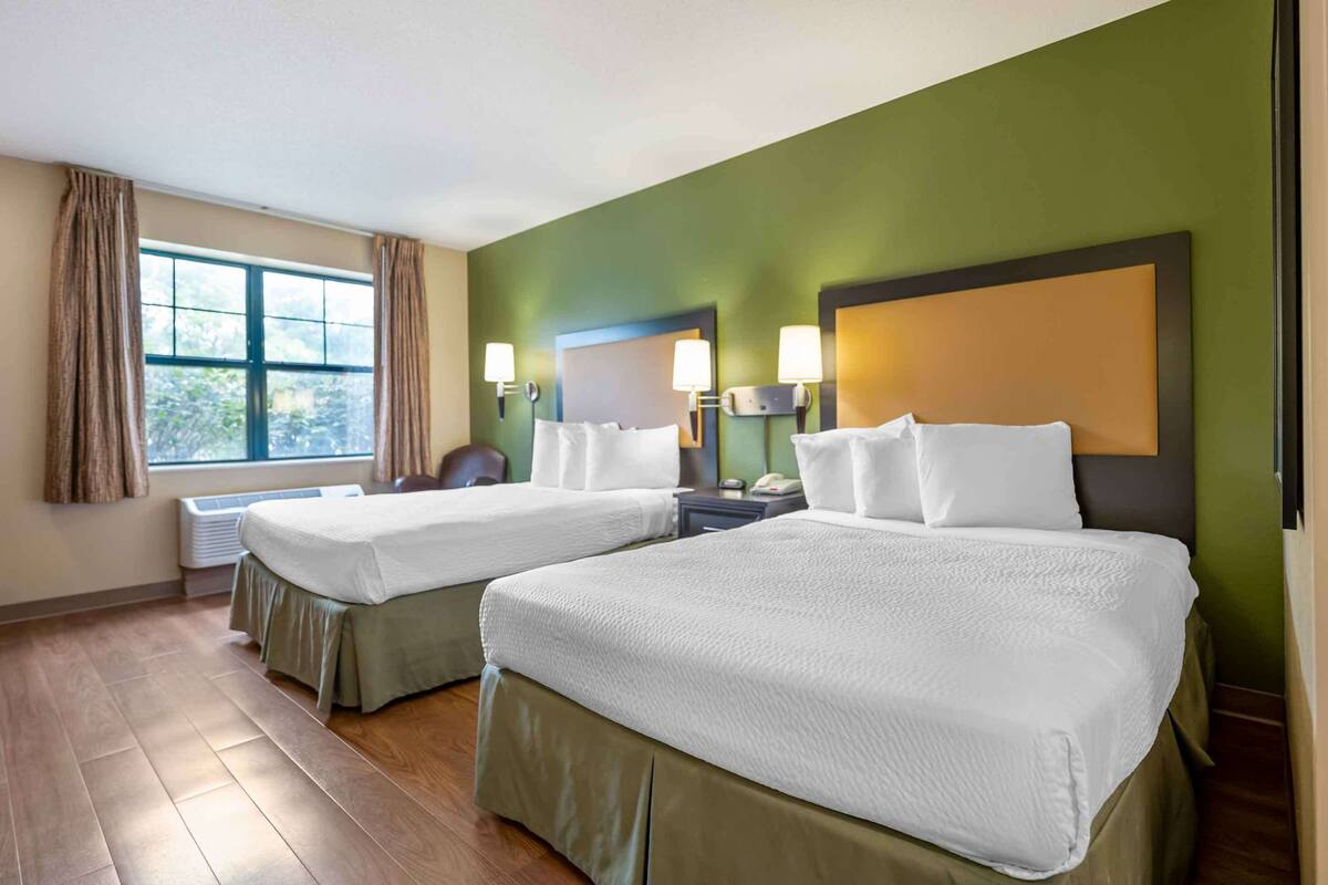 A hotel room with two beds, a window with curtains, a nightstand with a lamp, and a wall-mounted telephone.