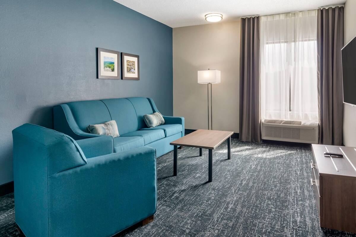 A modern hotel room with a teal sofa, a small wooden table, a floor lamp, and a large window with curtains.