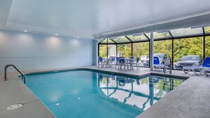 An indoor swimming pool with clear blue water, surrounded by blue and white lounge chairs, and large windows offering a view of parked cars and greenery outside. - Comfort Inn & Suites Troutville-Roanoke North (Troutville)
