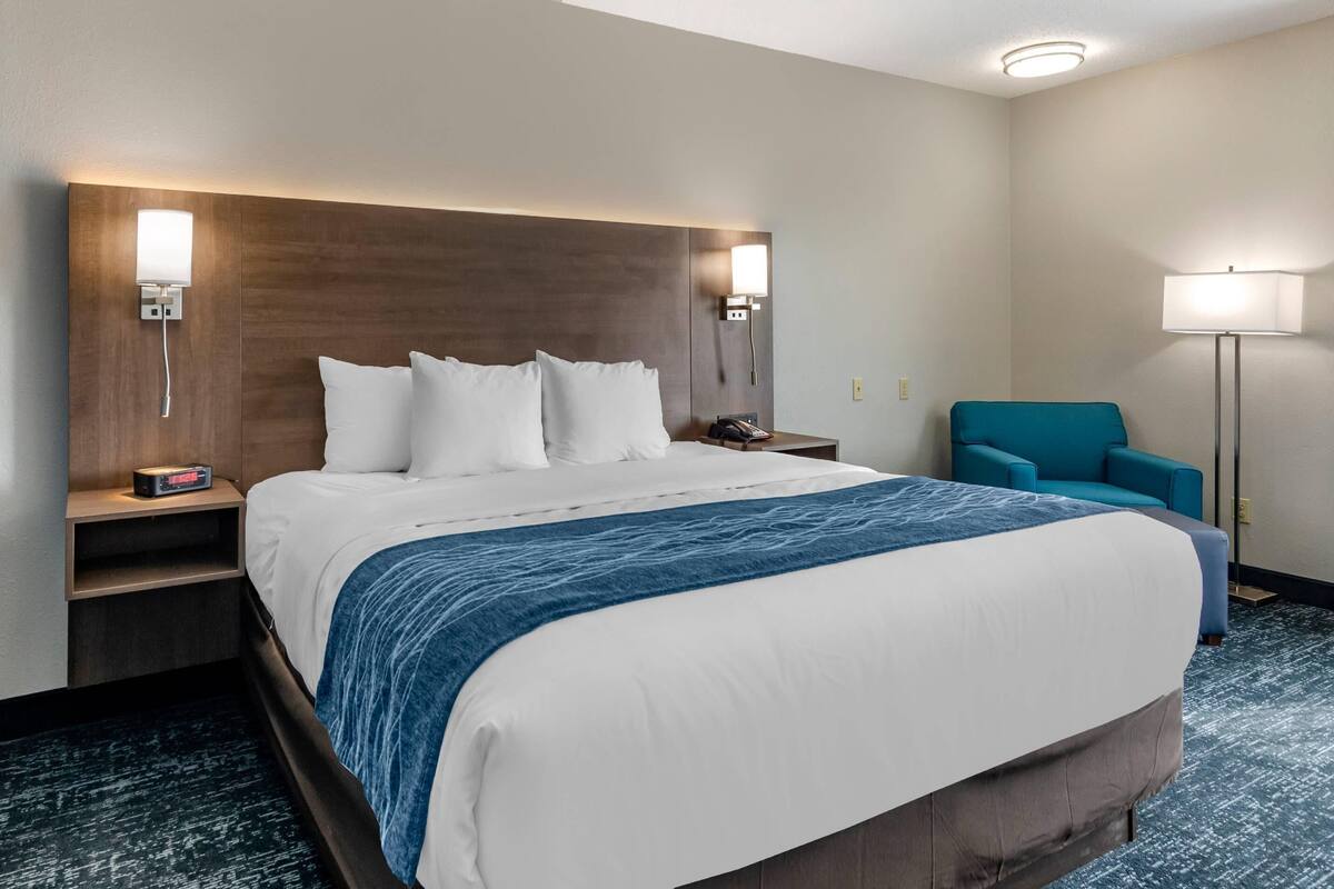 A hotel room with a large bed, a nightstand with a lamp, a chair, and a wall-mounted light.