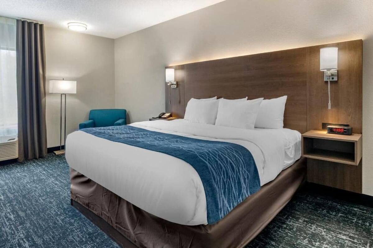 A hotel room with a large bed, a desk, a chair, a lamp, and a nightstand with a clock.