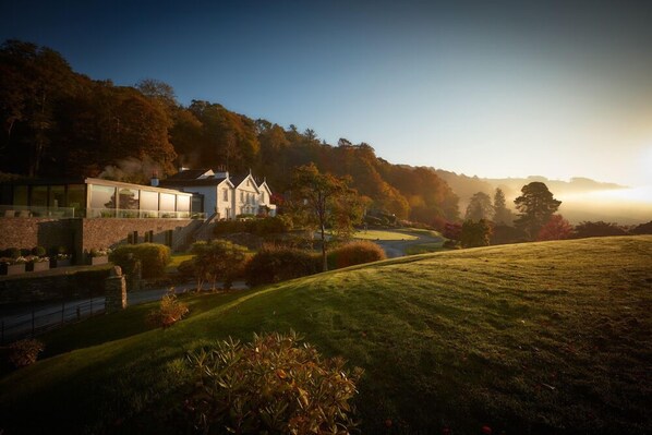A modern house with a large glass facade, surrounded by a well-maintained lawn and trees. - The Samling (Windermere)