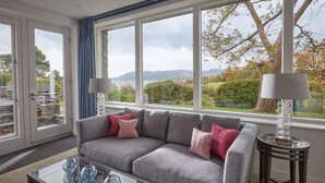 Ett modernt vardagsrum med en grå soffa, rosa kuddar, ett soffbord i glas och utsikt över en sjö och kullar.