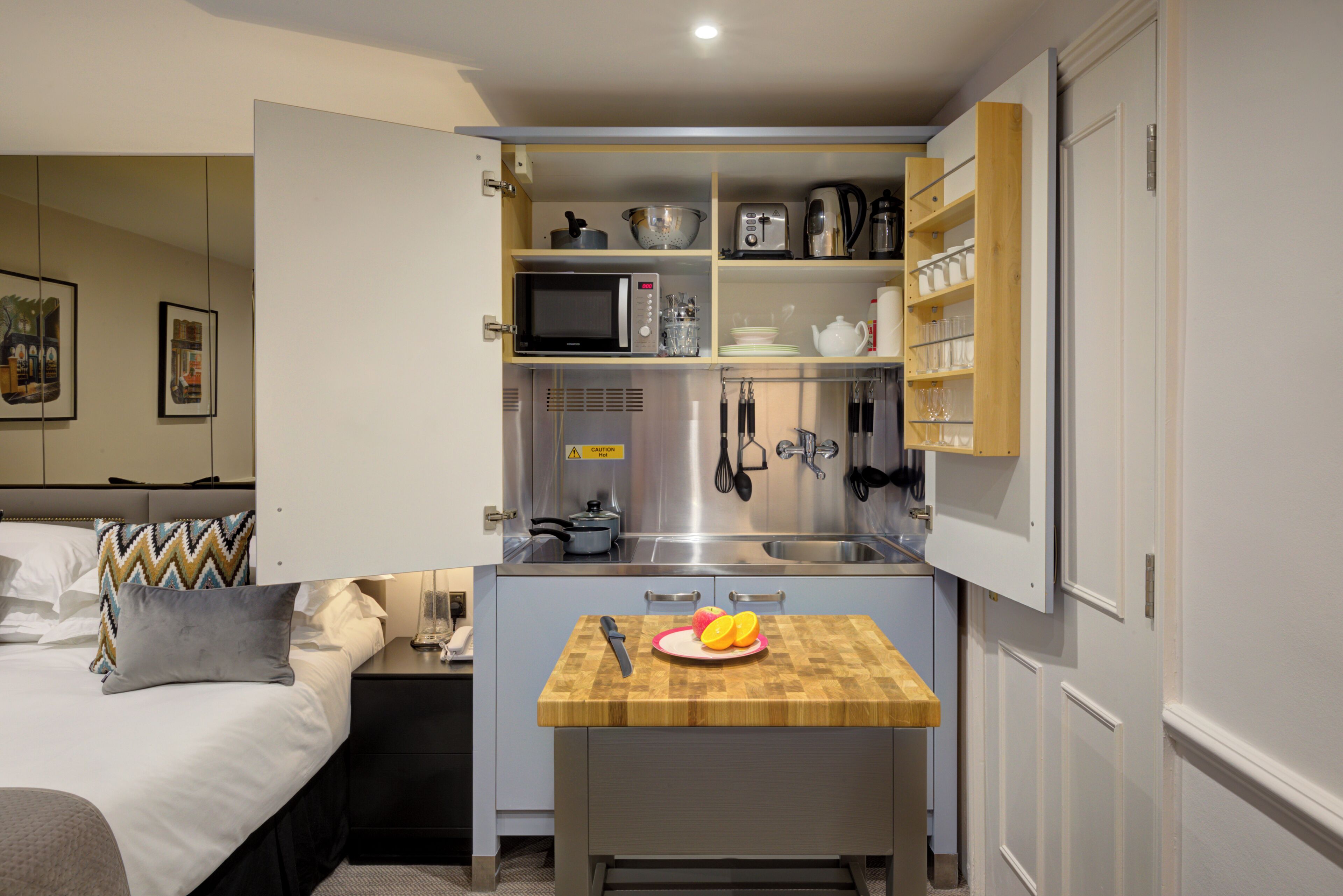 A compact kitchenette with a microwave, toaster, and sink, a small wooden table with fruit, and a bed with a patterned pillow.
