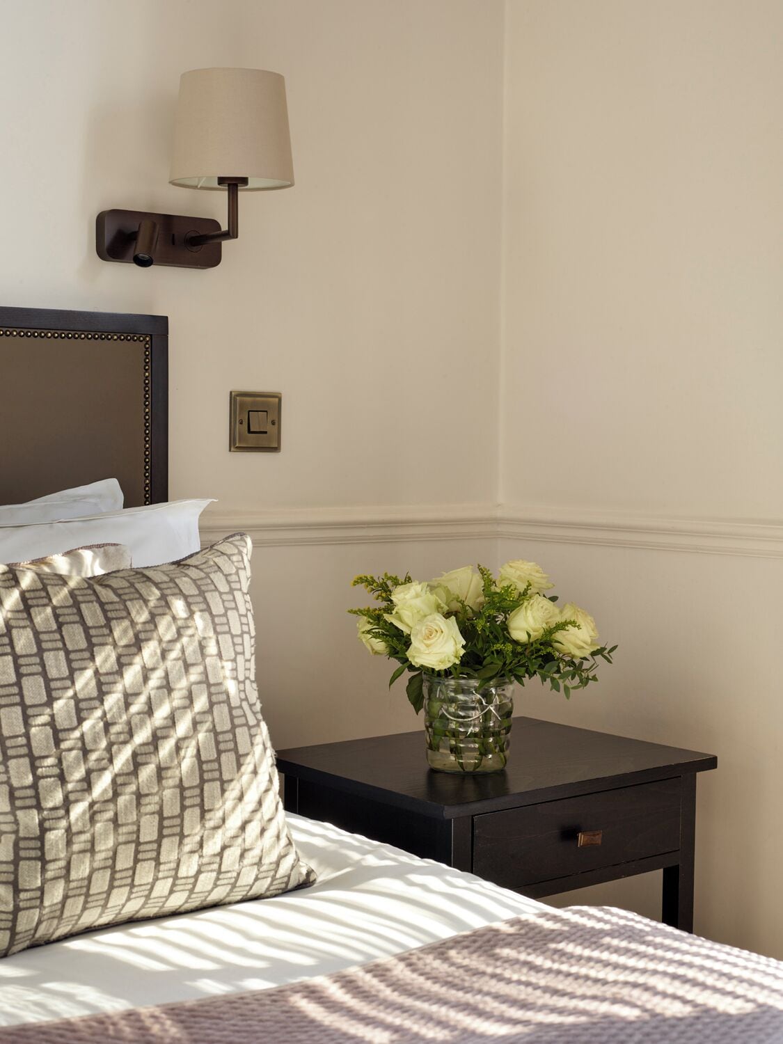 A neatly made bed with a patterned pillow, a bedside table with a vase of flowers, a wall-mounted lamp, and a light switch.