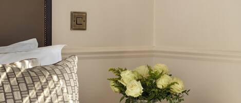 A neatly made bed with a patterned pillow, a bedside table with a vase of flowers, a wall-mounted lamp, and a light switch.