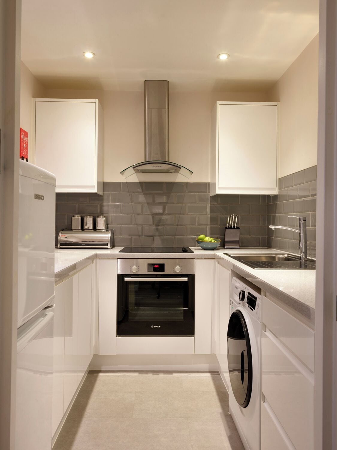 Une cuisine moderne avec des meubles blancs, un four Bosch, un lave-linge encastré et un dosseret en carrelage gris.
