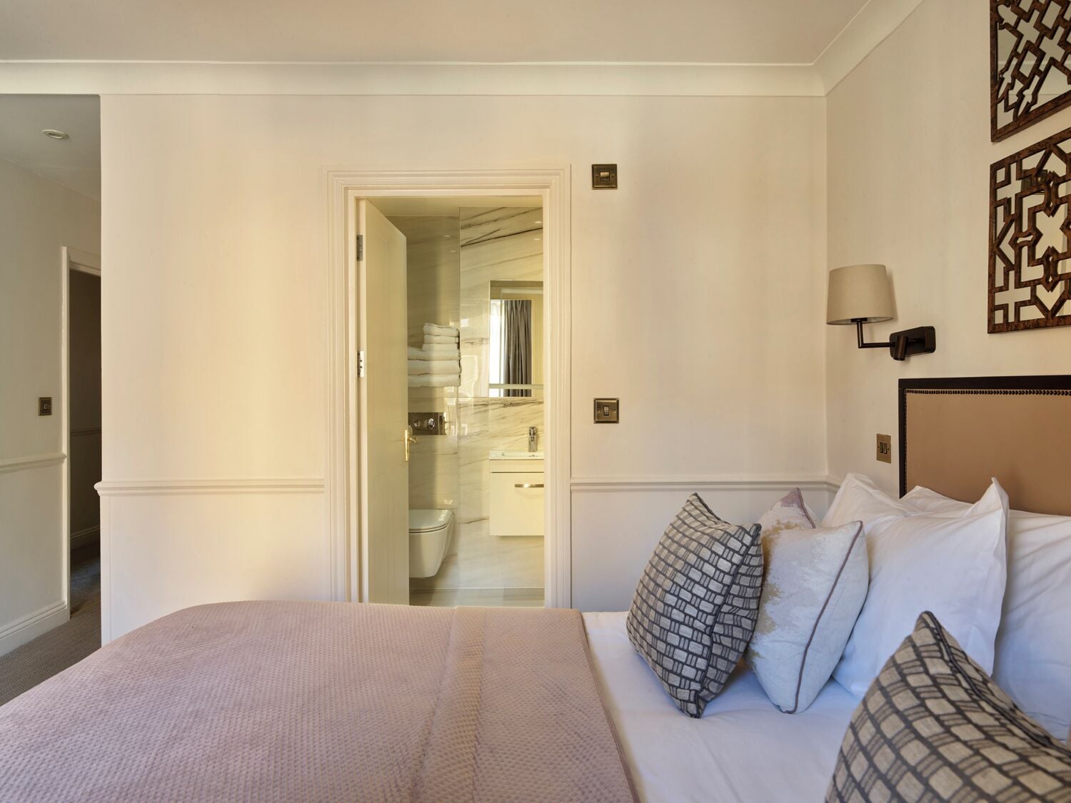 Une chambre d’hôtel avec un lit, une table de chevet, une lampe fixée au mur et une salle de bain visible à travers une porte ouverte.