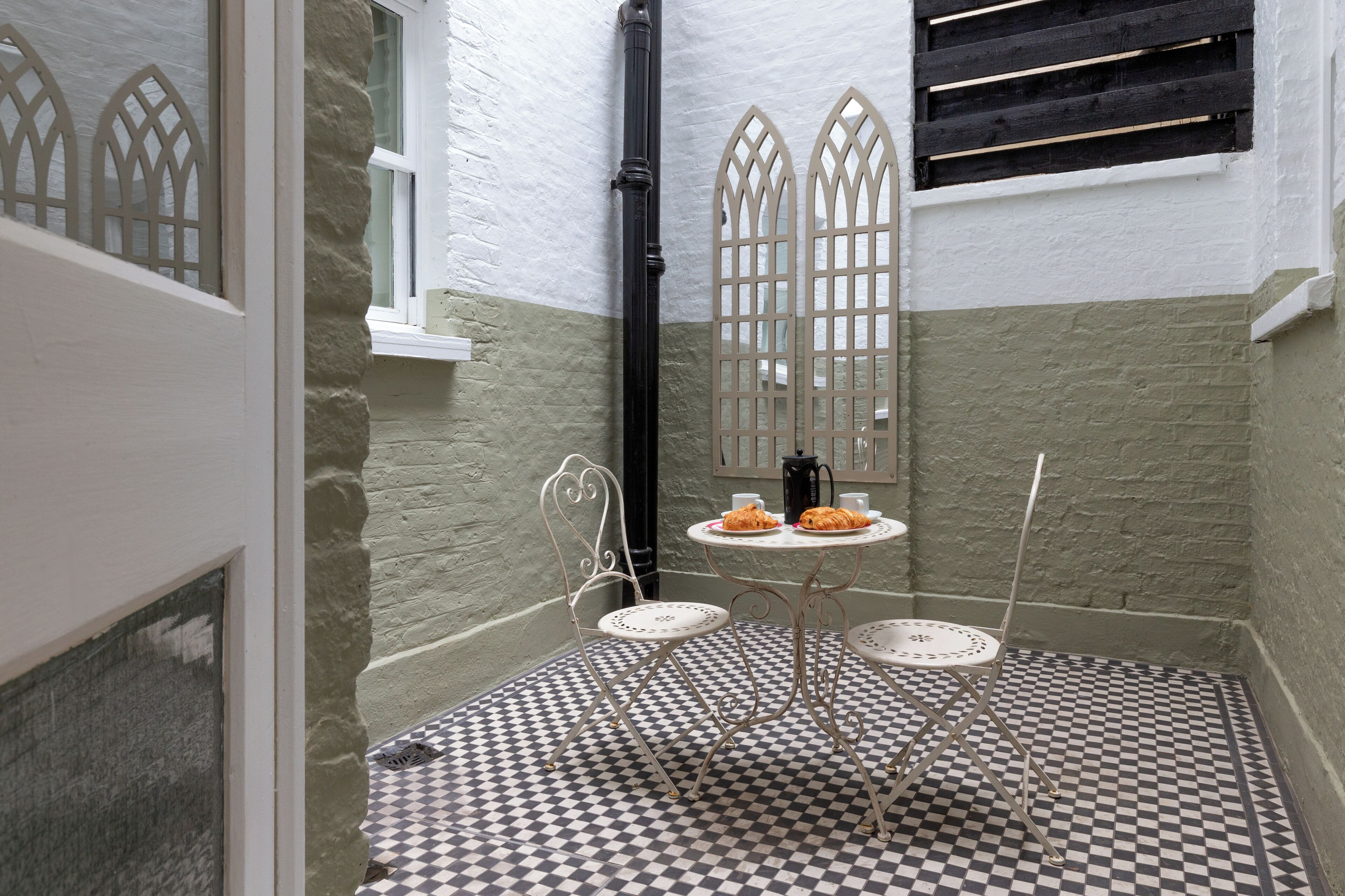A small room with a checkered floor, a white chair, a small table with a cup and pastries, and a window with a black shutter.