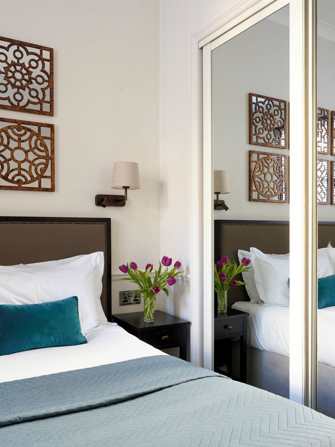 Ein Hotelzimmer mit Bett, Nachttisch mit Lampe, Blumenvase und dekorativen Wandpaneelen.