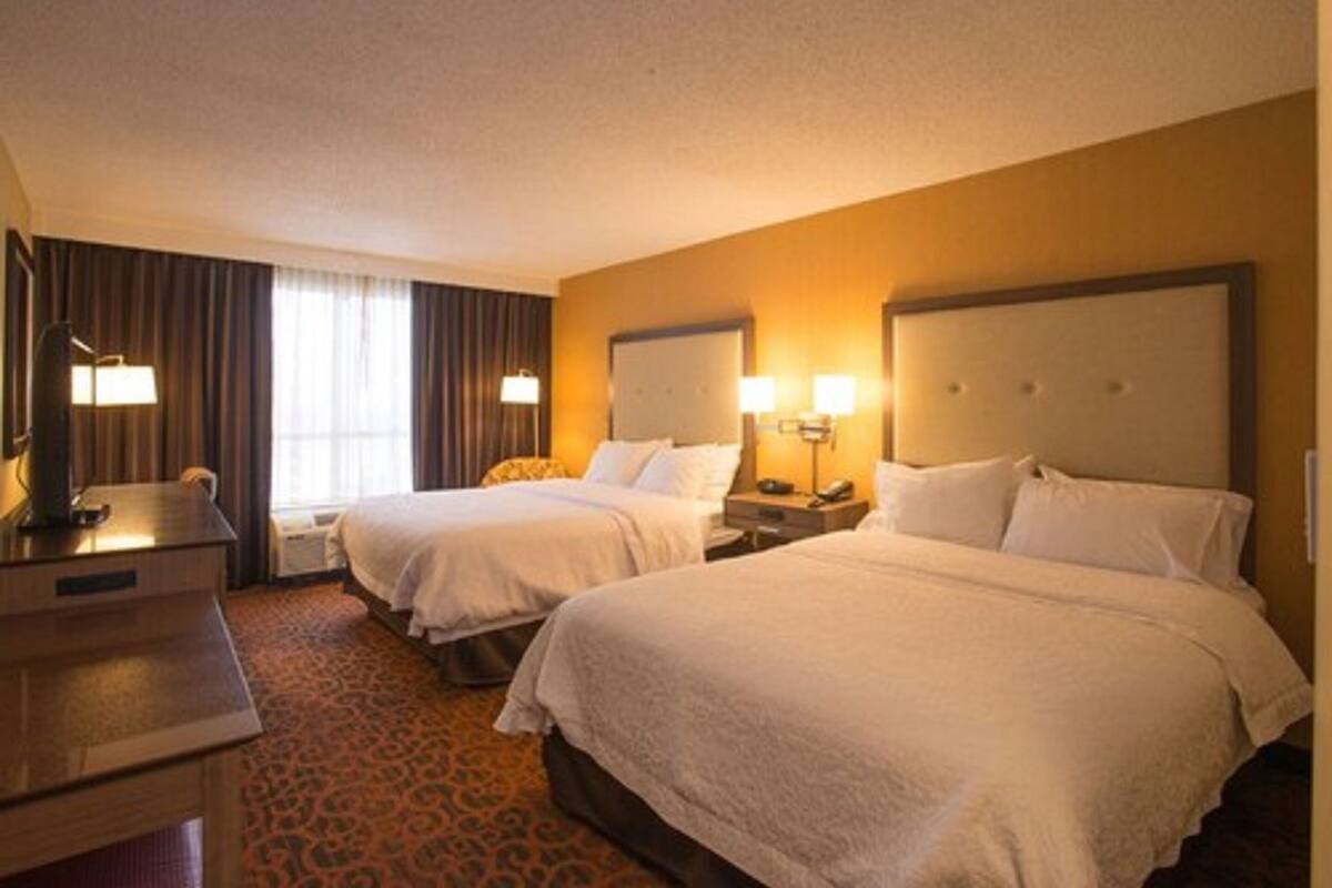 A hotel room with two beds, a desk, a lamp, and a window with curtains.