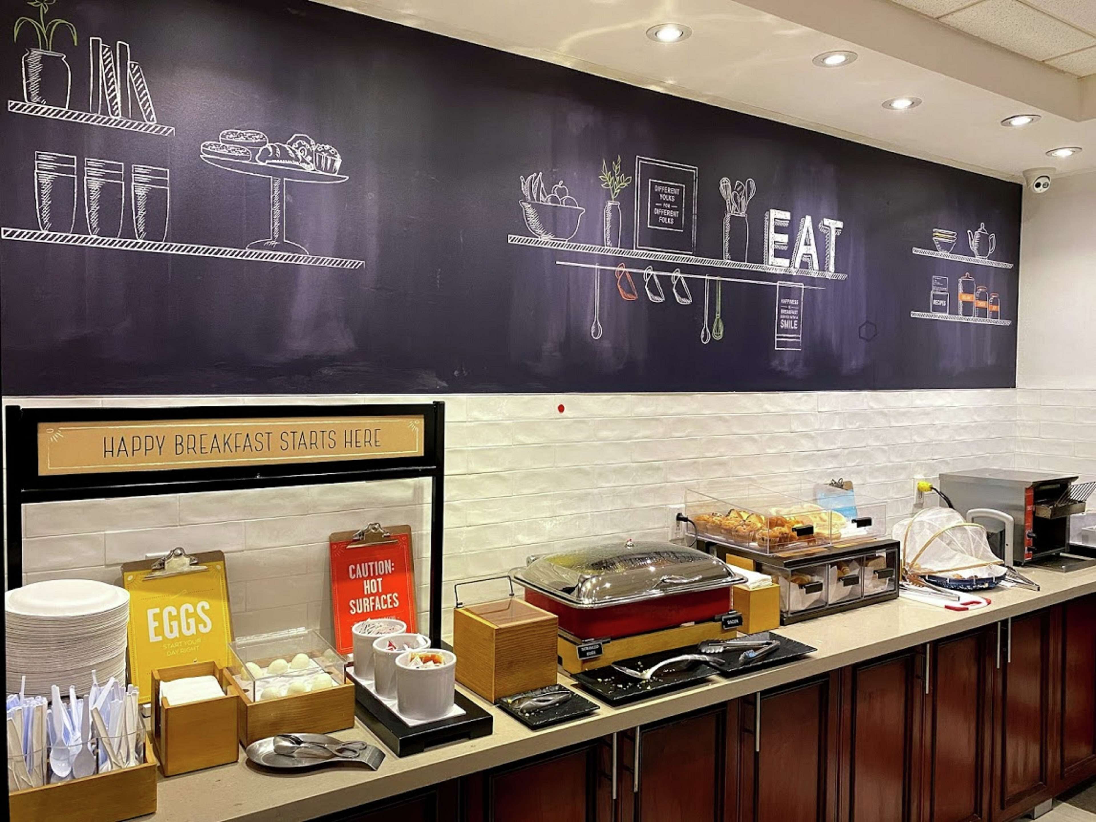 A breakfast buffet area with a chalkboard menu, a sign indicating hot surfaces, and various food and drink options.