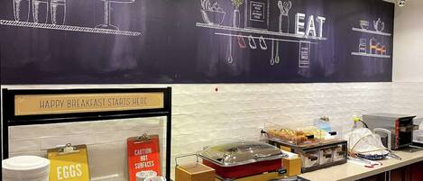 A breakfast buffet area with a chalkboard menu, a sign indicating hot surfaces, and various food and drink options.