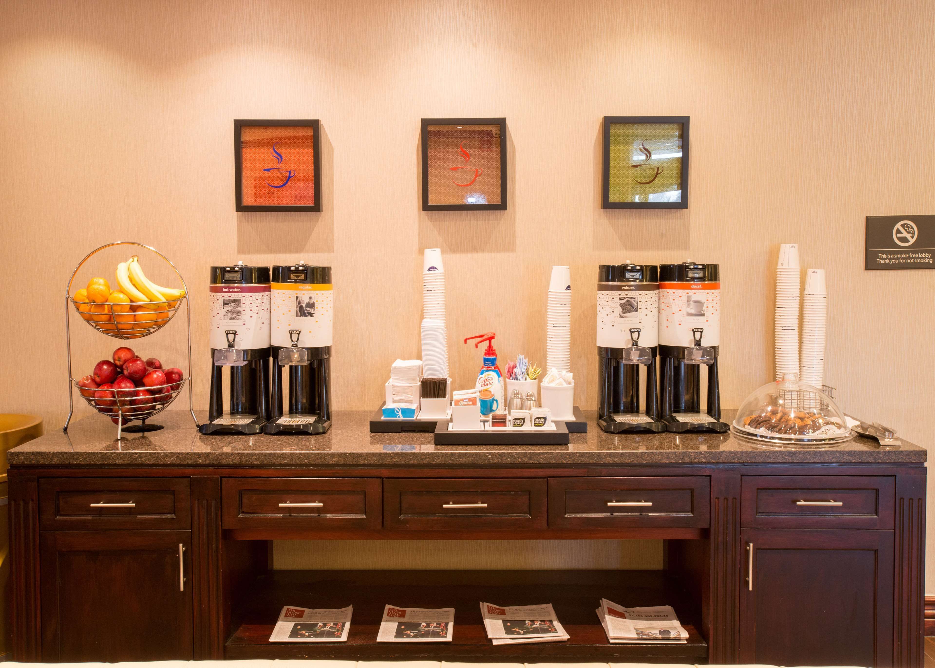 A hotel reception area with a fruit bowl, coffee and tea dispensers, and a stack of white cups.
