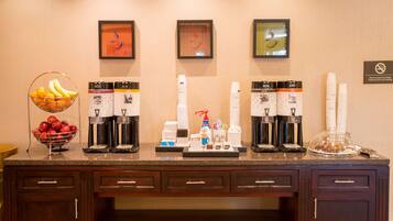A hotel reception area with a fruit bowl, coffee and tea dispensers, and a stack of white cups.