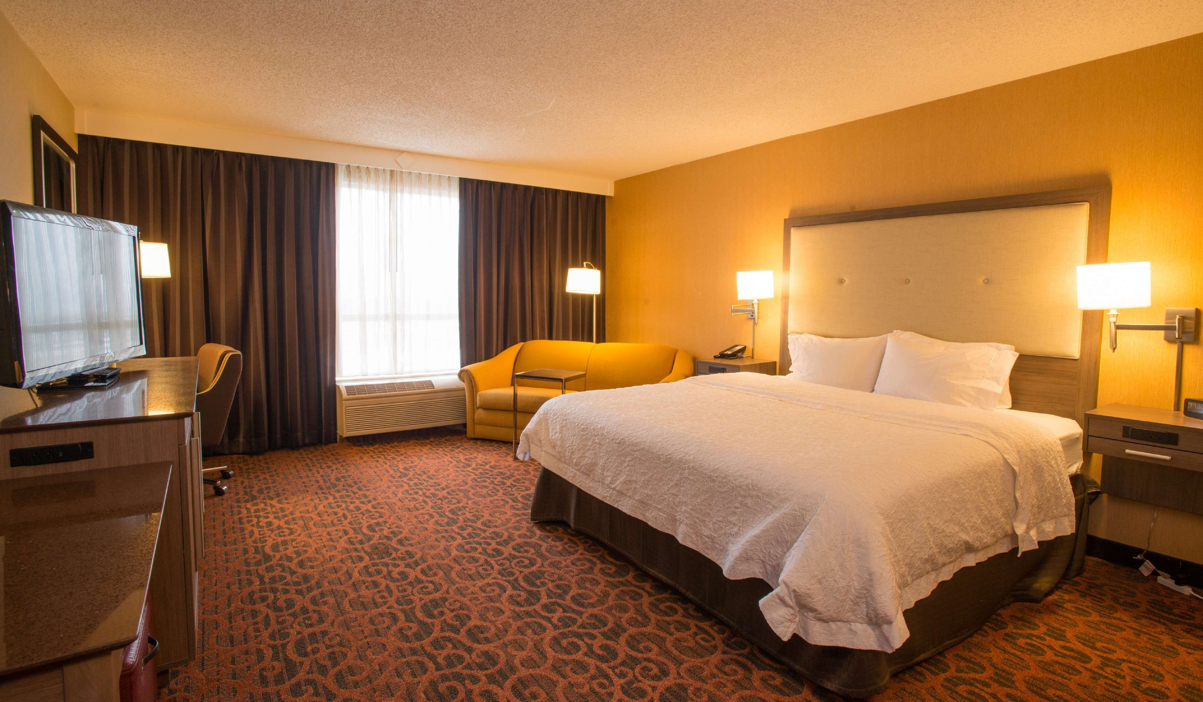 Ein Hotelzimmer mit einem großen Bett, einem Schreibtisch mit Fernseher, einem Sessel und einer Schreibtischlampe.