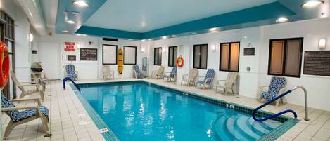 An indoor swimming pool with clear blue water, surrounded by lounge chairs and safety signs.