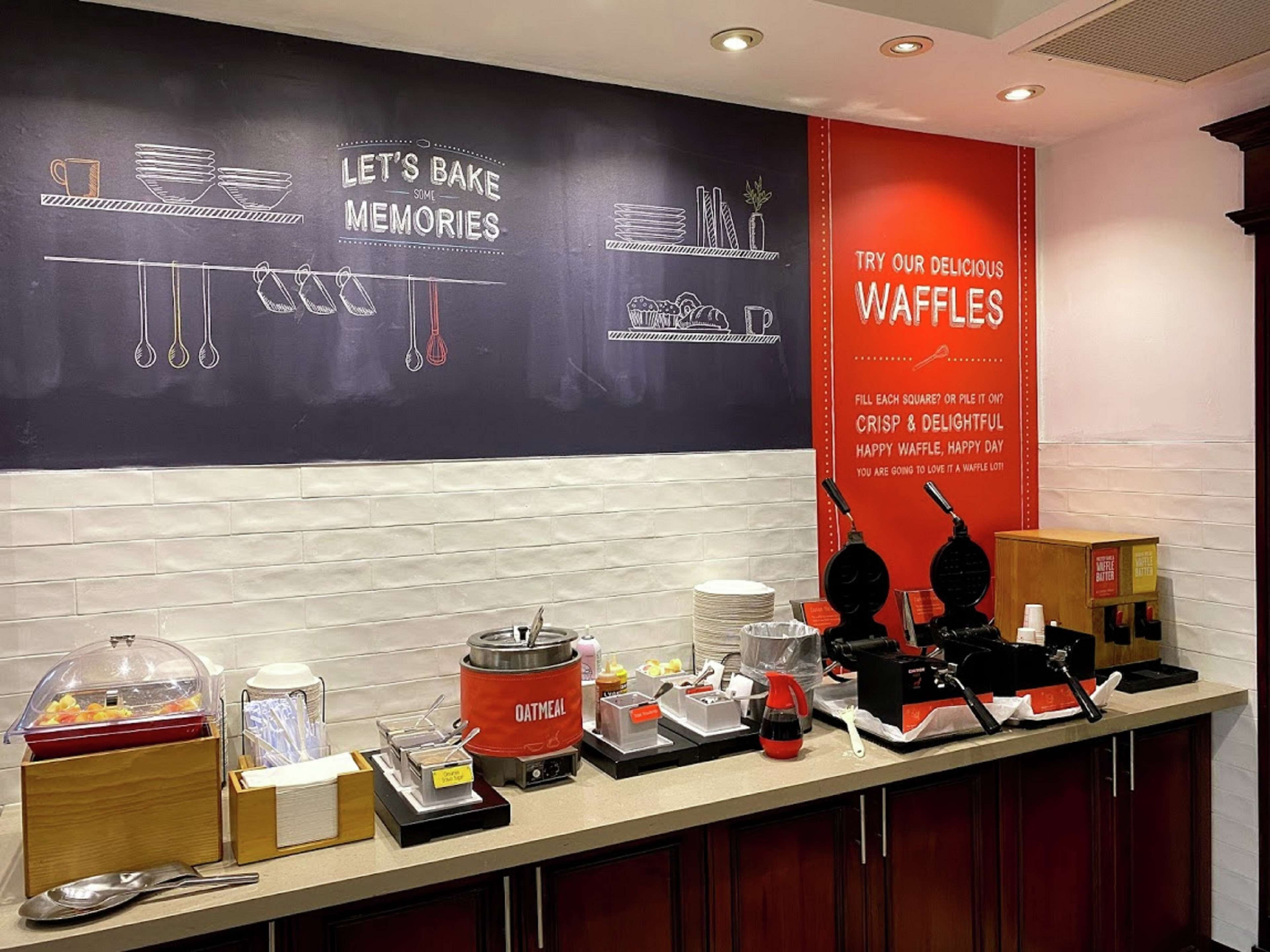 Zona de desayuno tipo bufé con una pizarra que muestra 'Let's Bake Some Memories' y un letrero rojo que anuncia waffles.