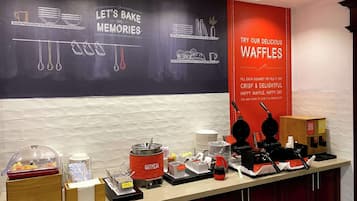 Zona de desayuno tipo bufé con una pizarra que muestra 'Let's Bake Some Memories' y un letrero rojo que anuncia waffles.