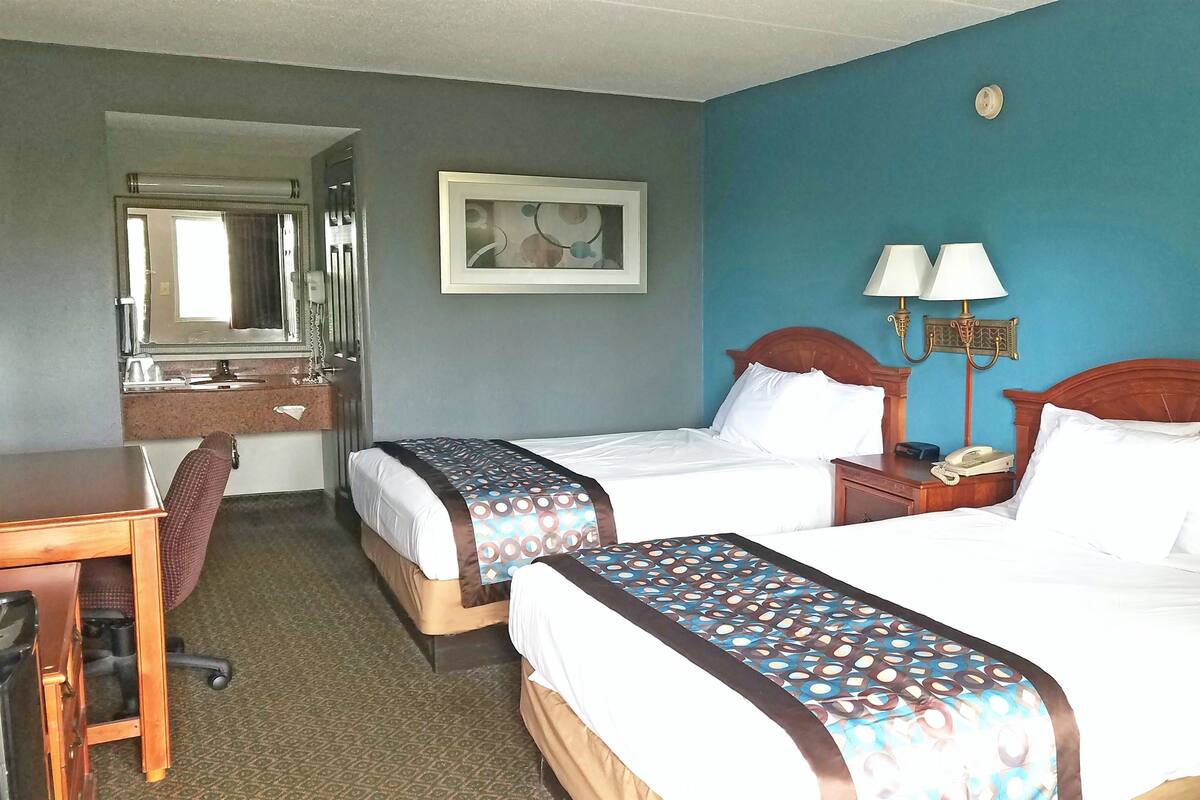 A hotel room with two beds, a desk, a chair, a mirror, and a wall-mounted lamp.