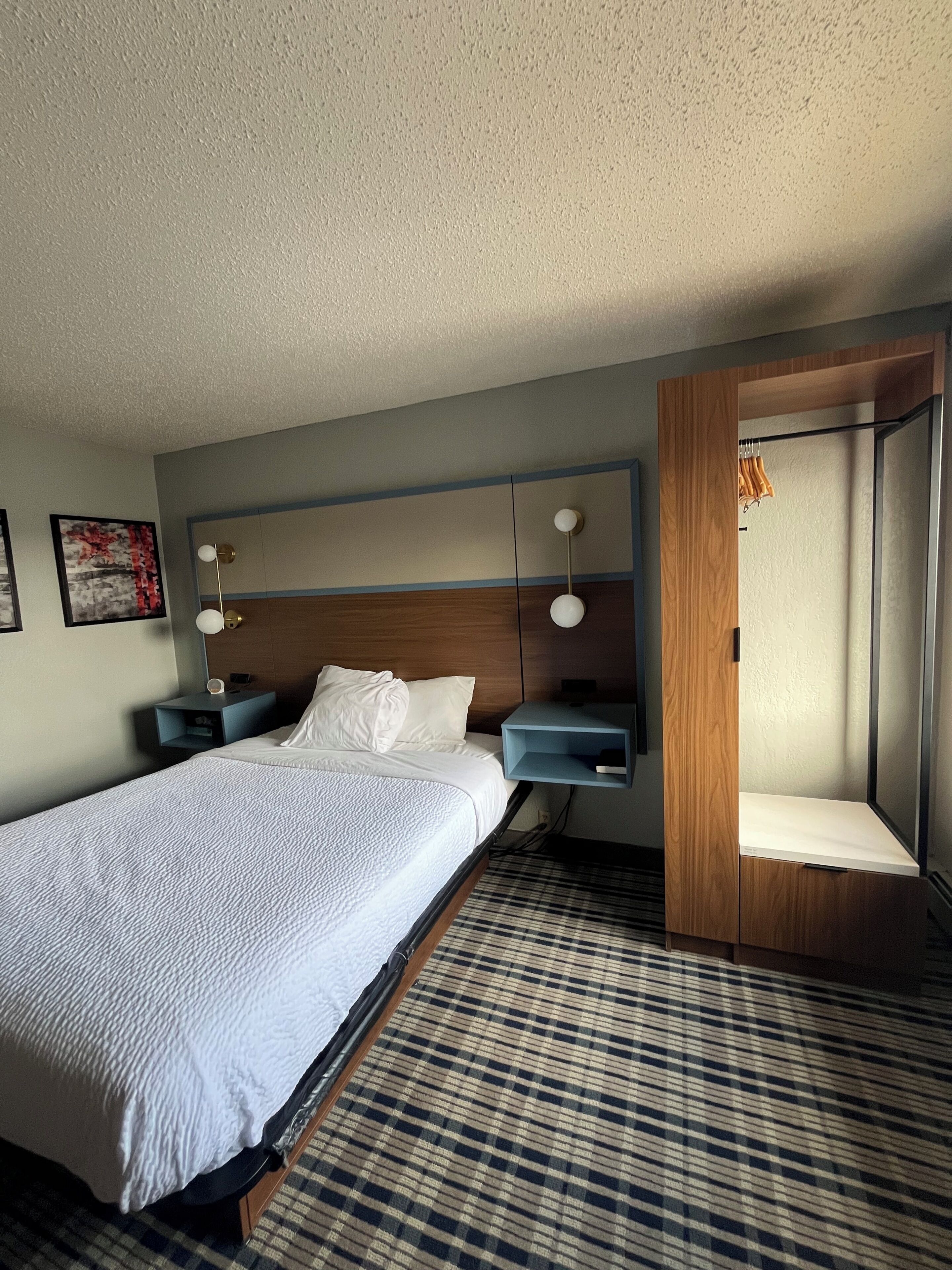 Habitación de hotel con una cama grande, una mesita de noche, un estante en la pared y una puerta con espejo.