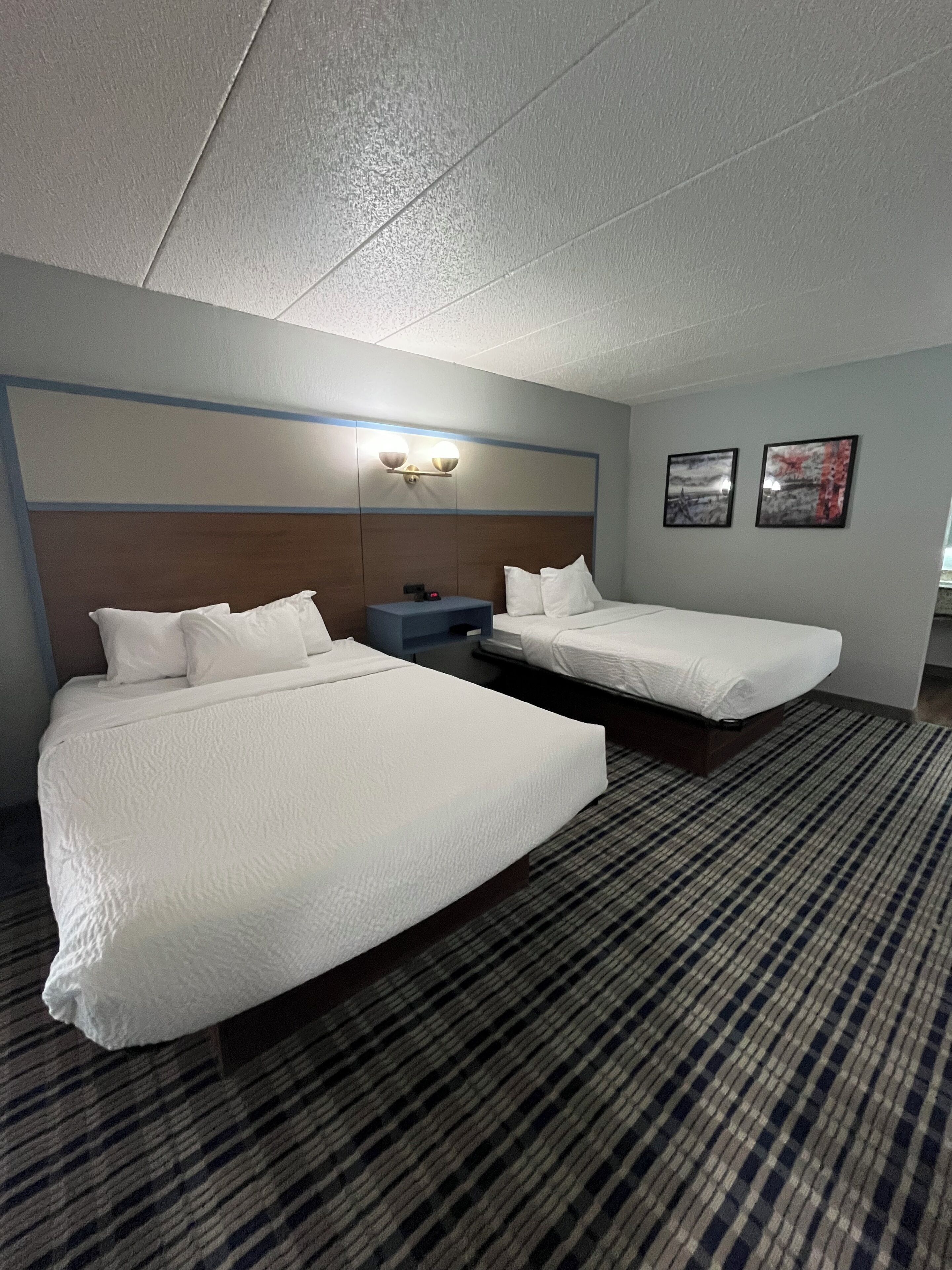Habitación de hotel con dos camas, una mesita de noche, una lámpara de pared y dos cuadros enmarcados.