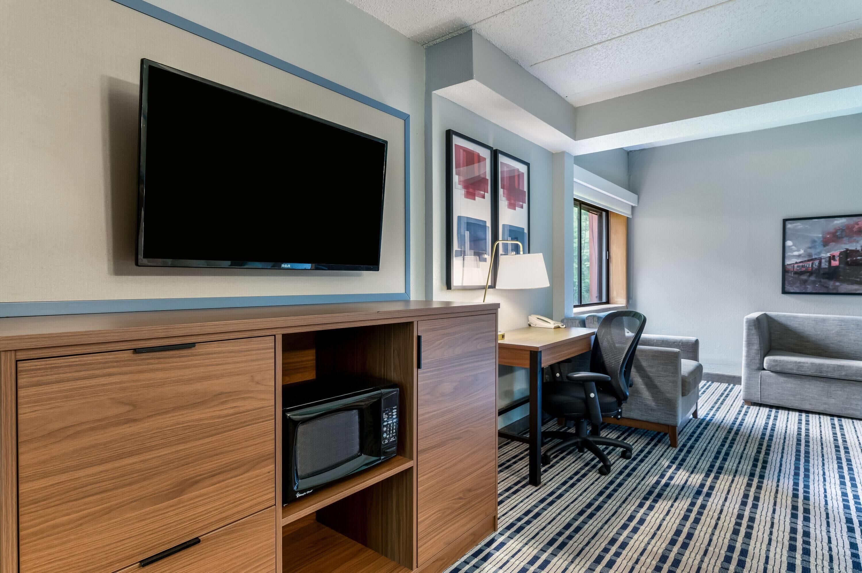 A hotel room with a flat-screen TV, a desk with a lamp, a microwave, a chair, and a couch.