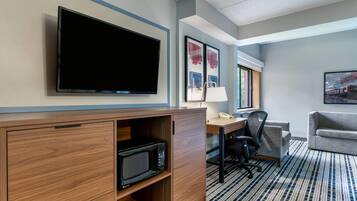 A hotel room with a flat-screen TV, a desk with a lamp, a microwave, a chair, and a couch.
