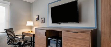 Een hotelkamer met een wandgemonteerde flatscreen televisie, een bureau met lamp, een stoel en een magnetron in een houten kast.