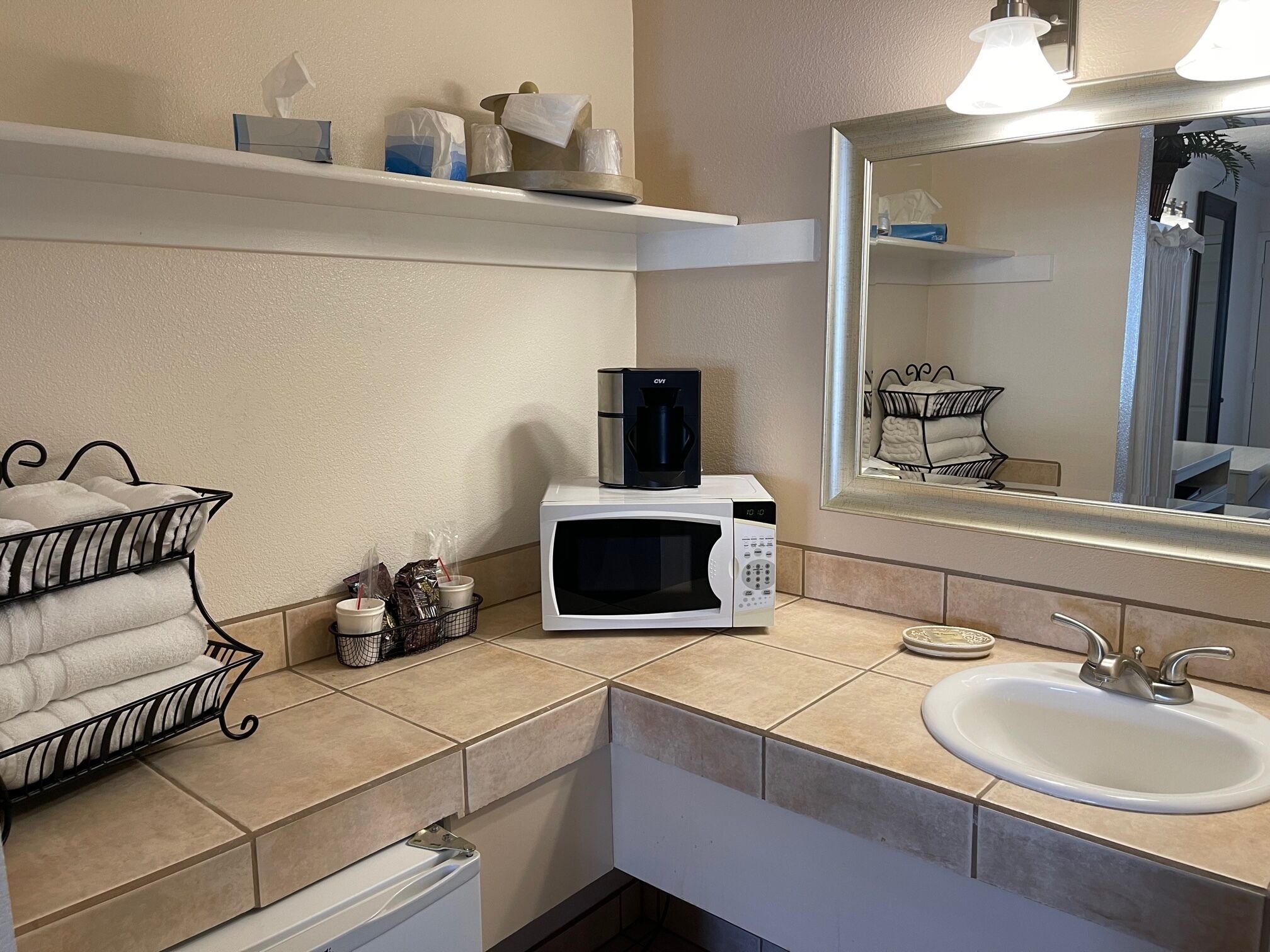 Un pequeño kitchenette con microondas, cafetera y lavamanos. Hay toallas y artículos de aseo en los estantes.