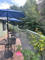 Un patio al aire libre con sillas de metal y una sombrilla azul, rodeado por una valla de madera y abundante vegetación.