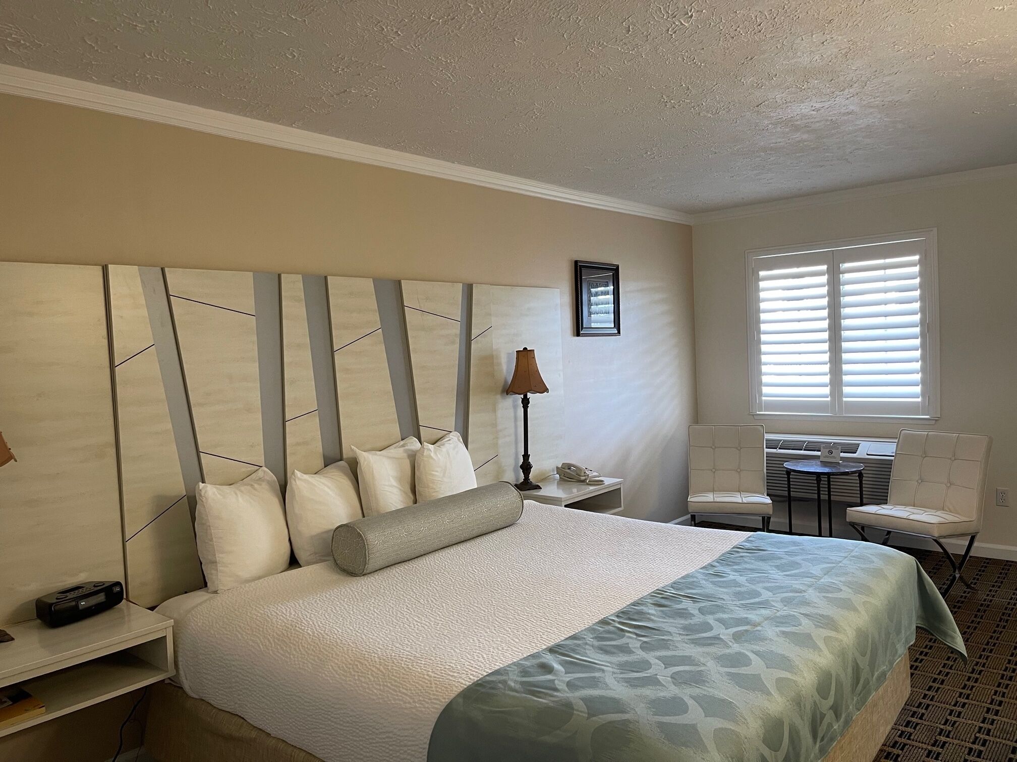 Habitación de hotel con una cama grande, un escritorio con silla, una lámpara y una ventana con persianas.