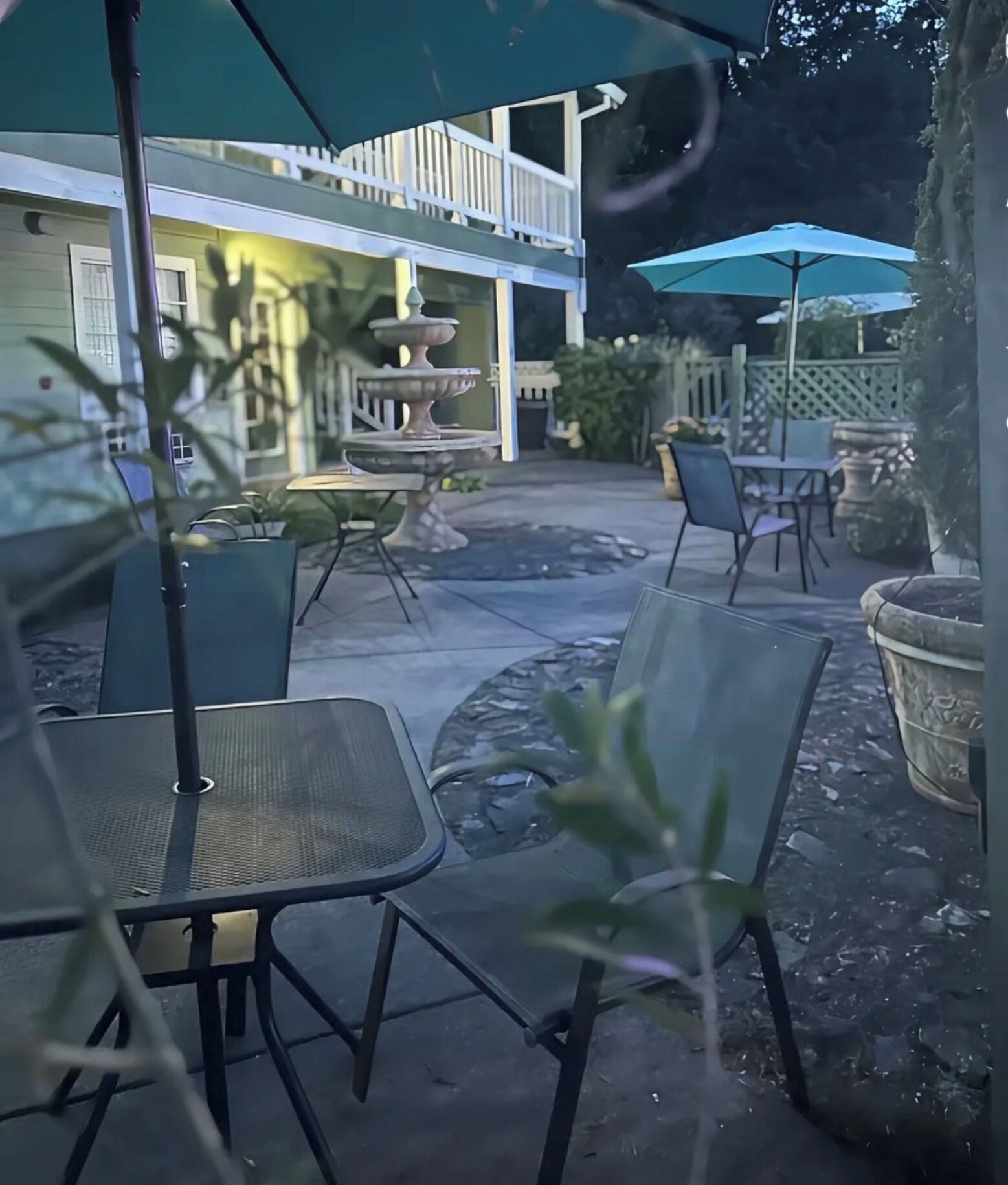 Un espacio al aire libre con patio, sillas y mesas, una fuente decorativa y un edificio con balcón al fondo.