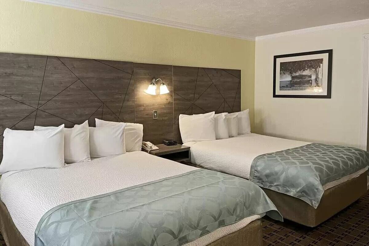 Quarto de hotel com duas camas, cabeceira com design geométrico, luminária fixada na parede e quadro na parede.