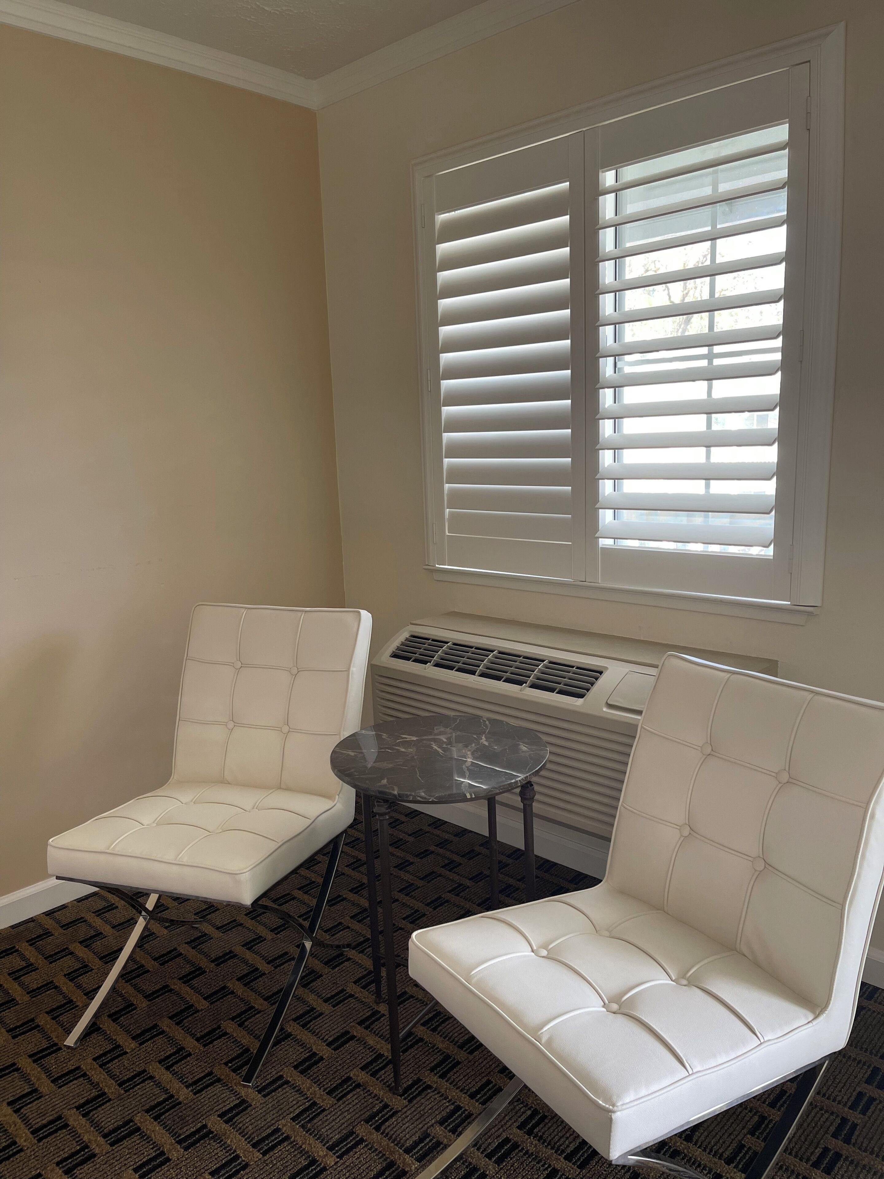 A room with two white cushioned chairs, a small round table, and a window with blinds.