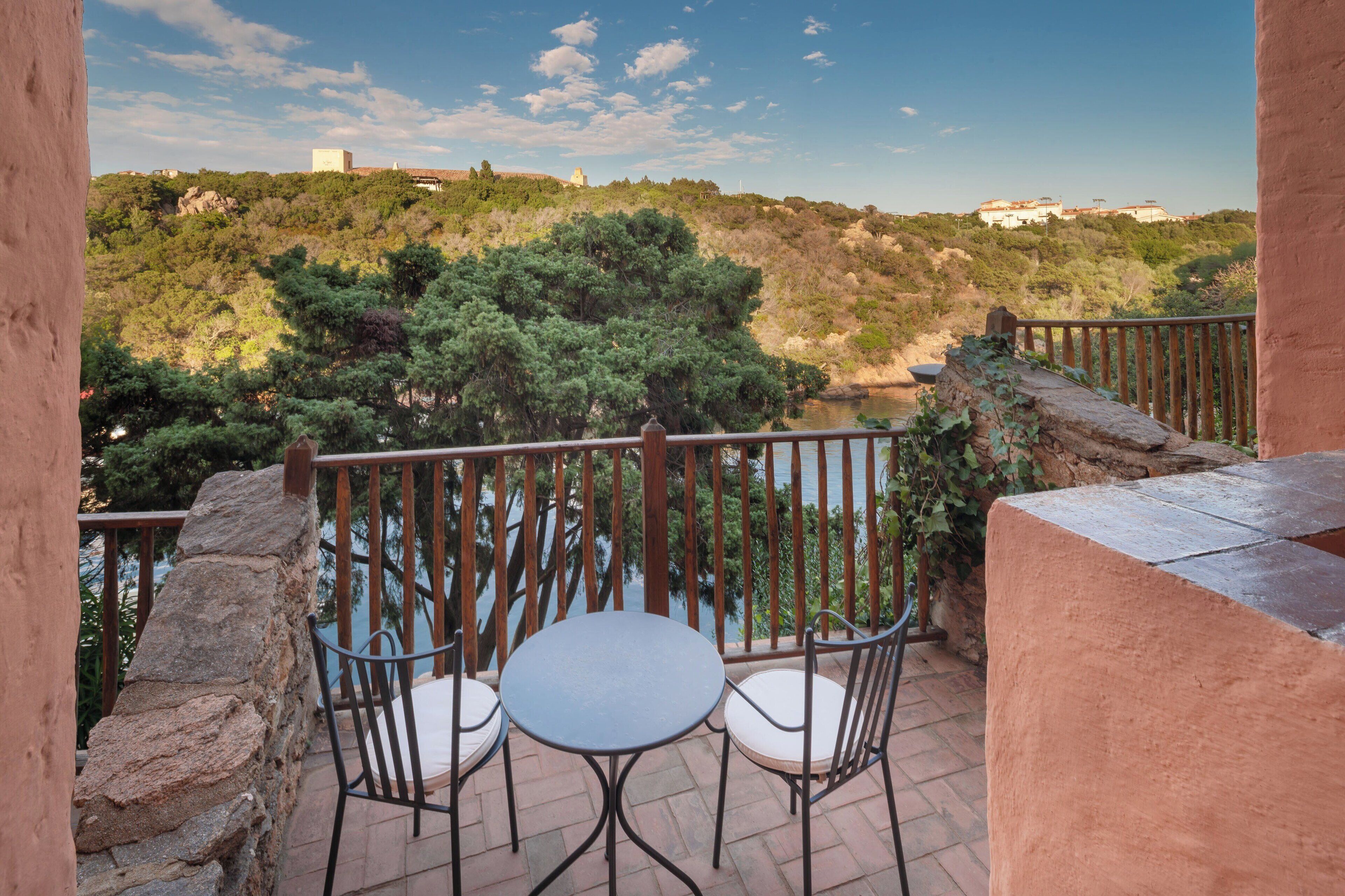 A balcony with two chairs and a table, overlooking a river and a hilly landscape with buildings in the distance.