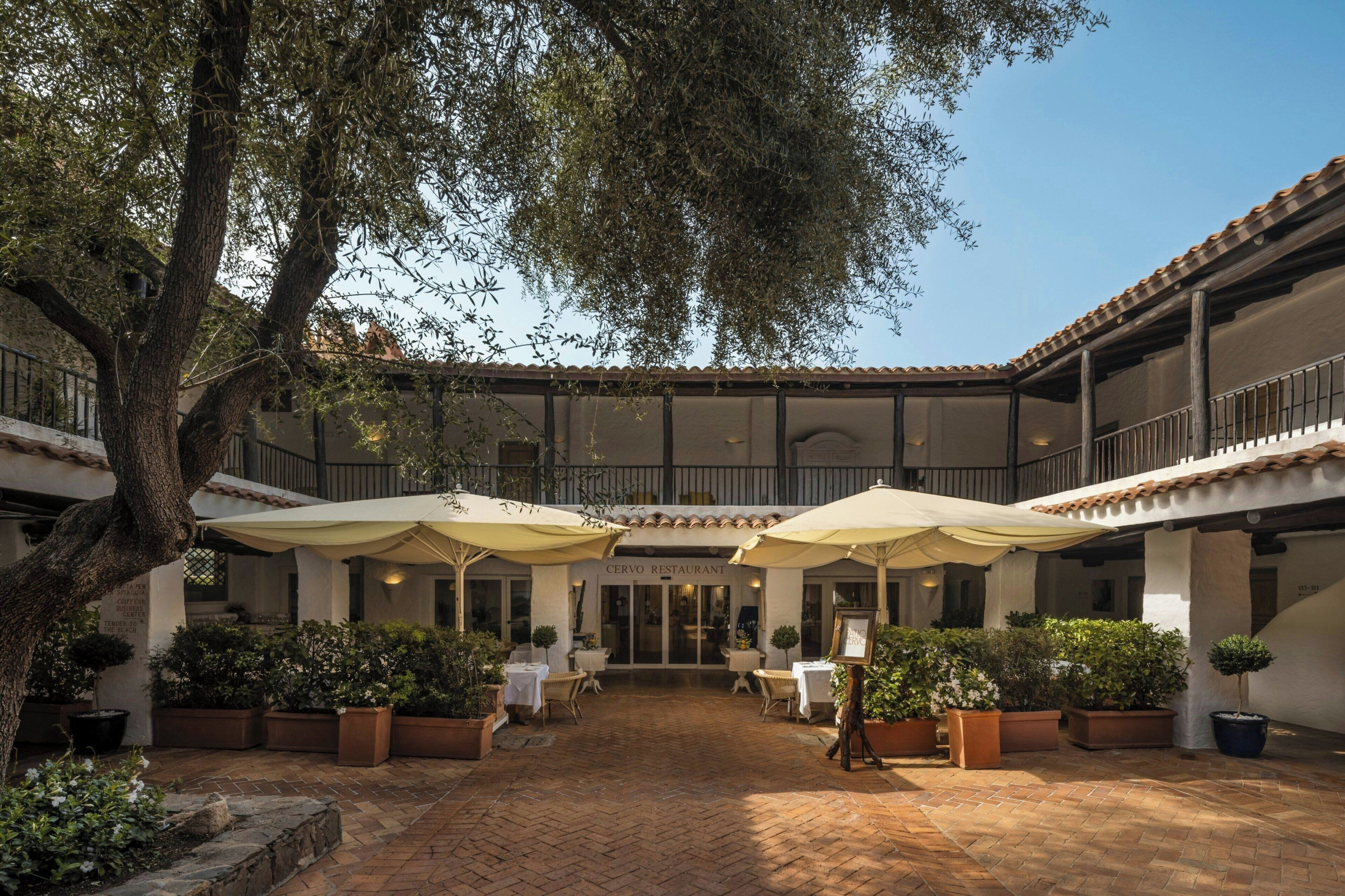 A courtyard with a restaurant named 'Cervo Restaurant' in the center, surrounded by potted plants and outdoor seating.