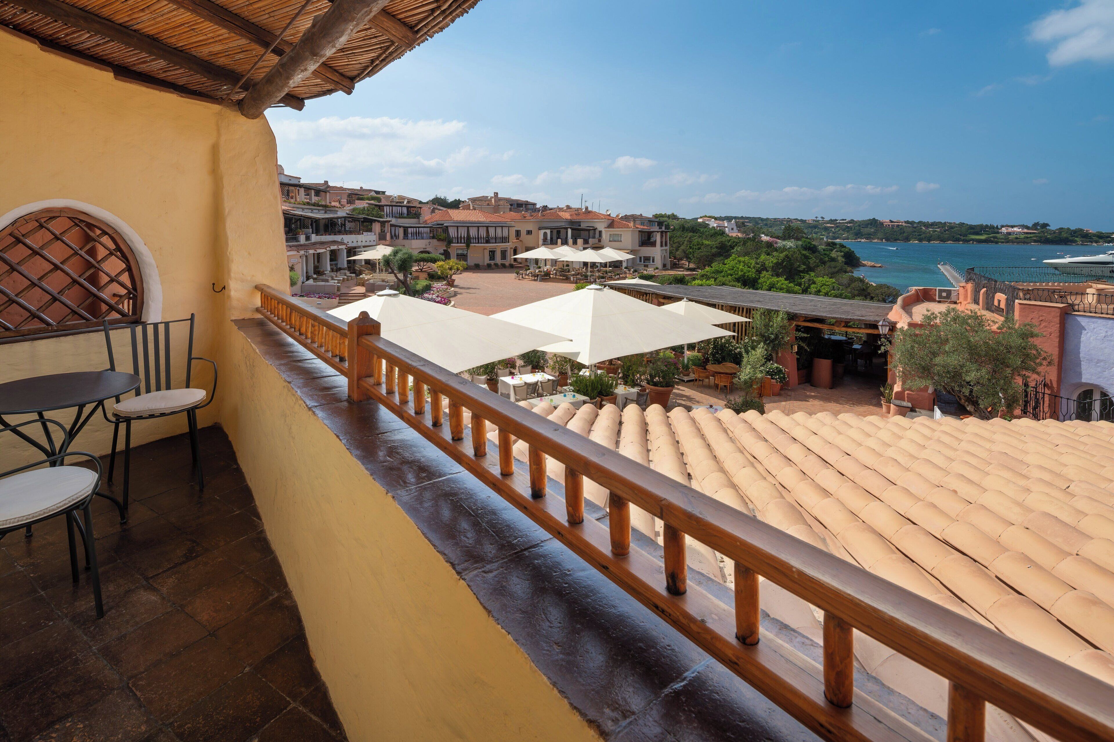 Un balcón con baranda de madera que da a una terraza con mobiliario exterior y sombrillas, vista al mar y edificios tradicionales al fondo.