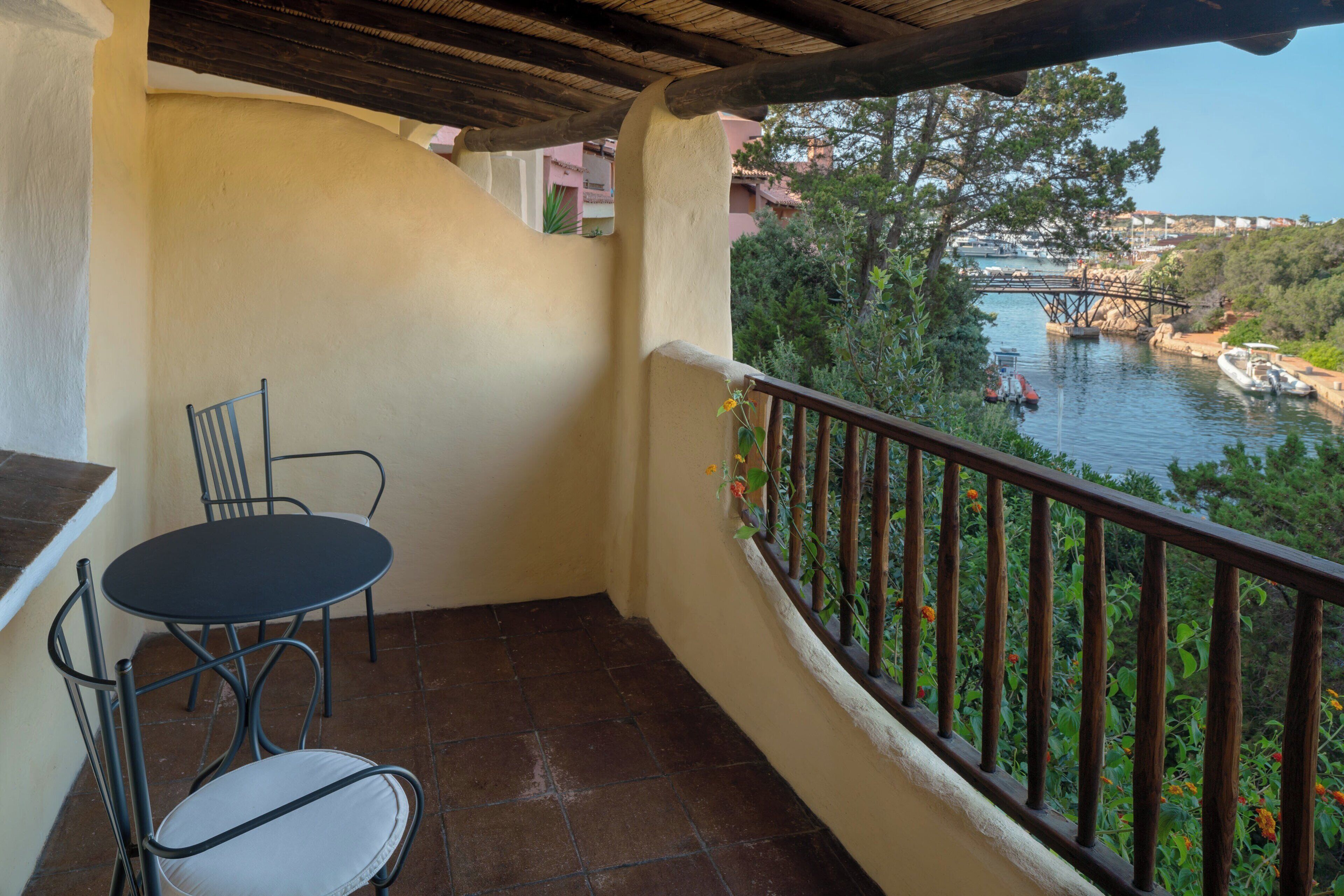 A balcony with a table and chairs, overlooking a river with boats and a bridge.