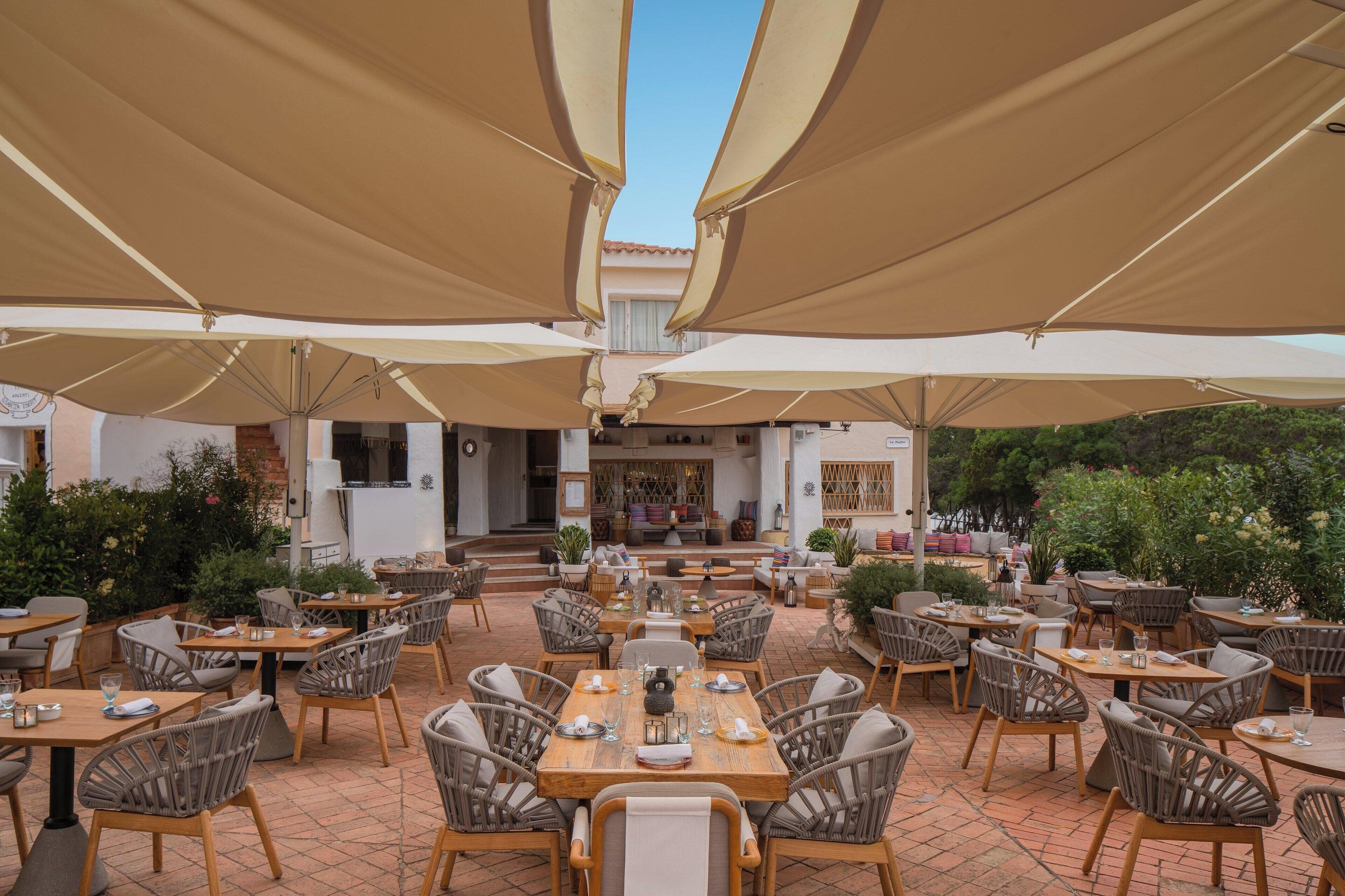 An outdoor dining area with multiple tables and chairs, shaded by large umbrellas, and a view of the interior through a glass door.