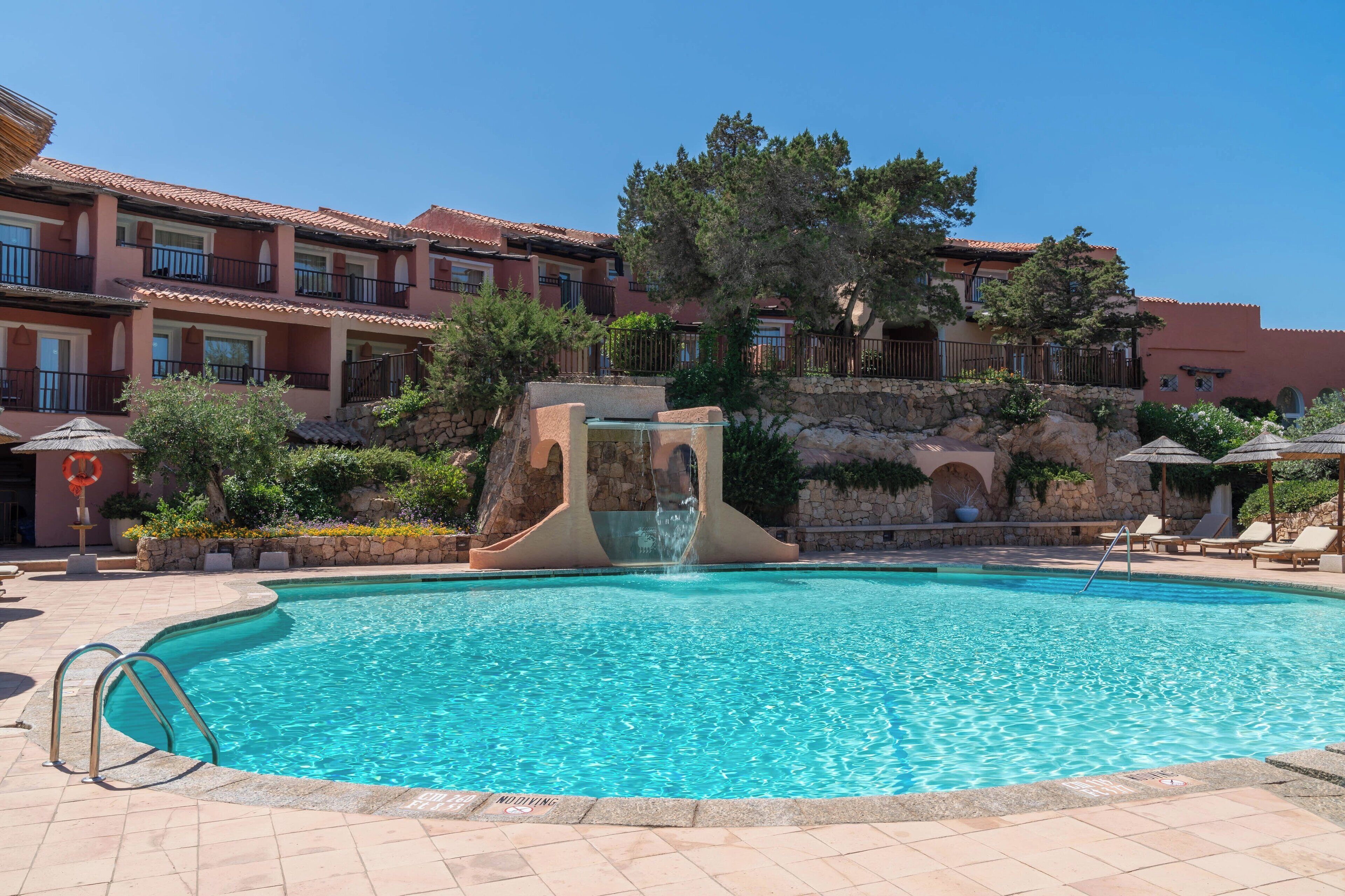Una piscina con fuente, rodeada por un muro de piedra y un edificio con balcones.