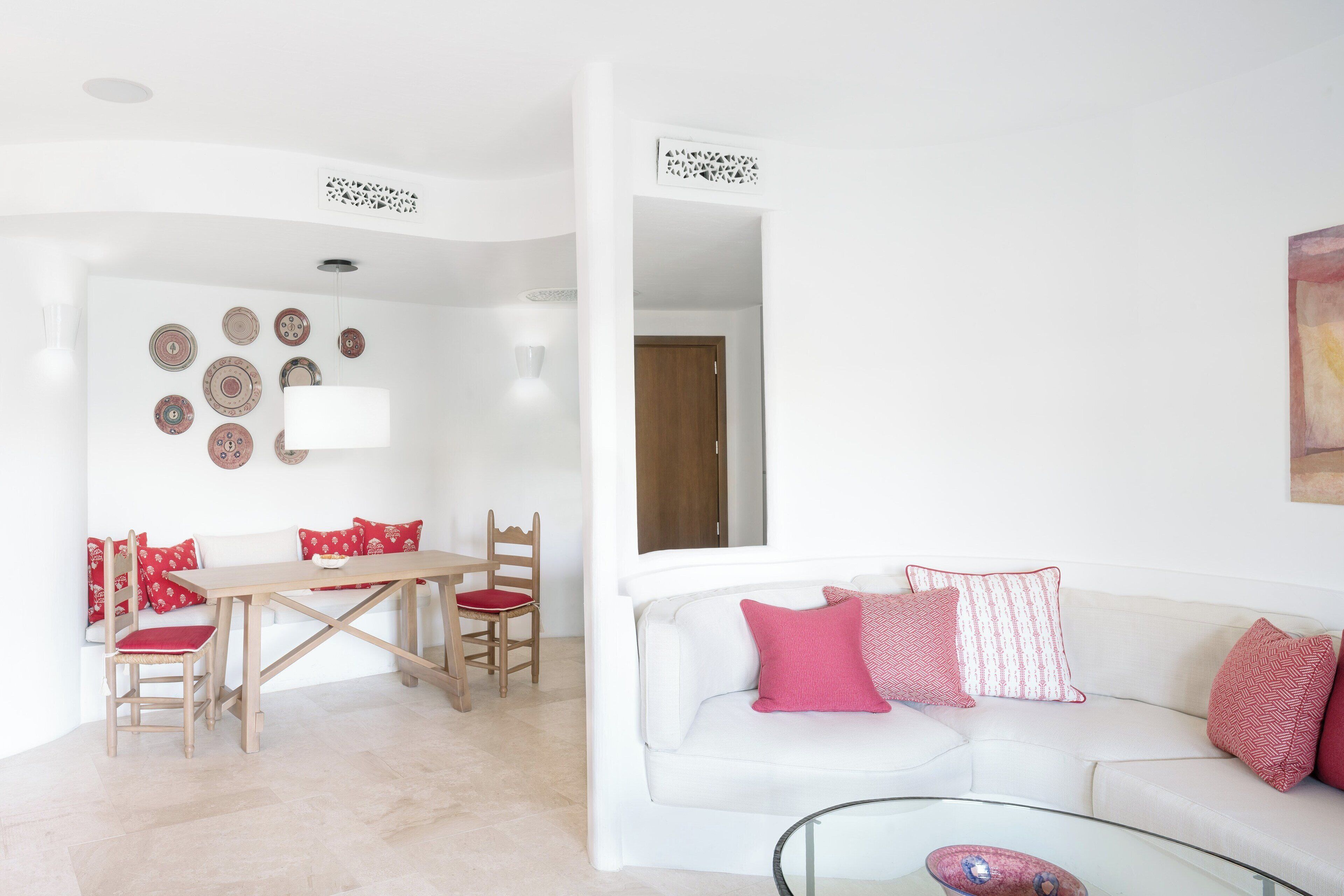 Un salón con un sofá blanco, cojines rojos, una mesa de centro de vidrio y una mesa de comedor de madera con sillas.