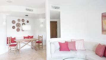 A living room with a white sofa, red cushions, a glass coffee table, and a wooden dining table with chairs.